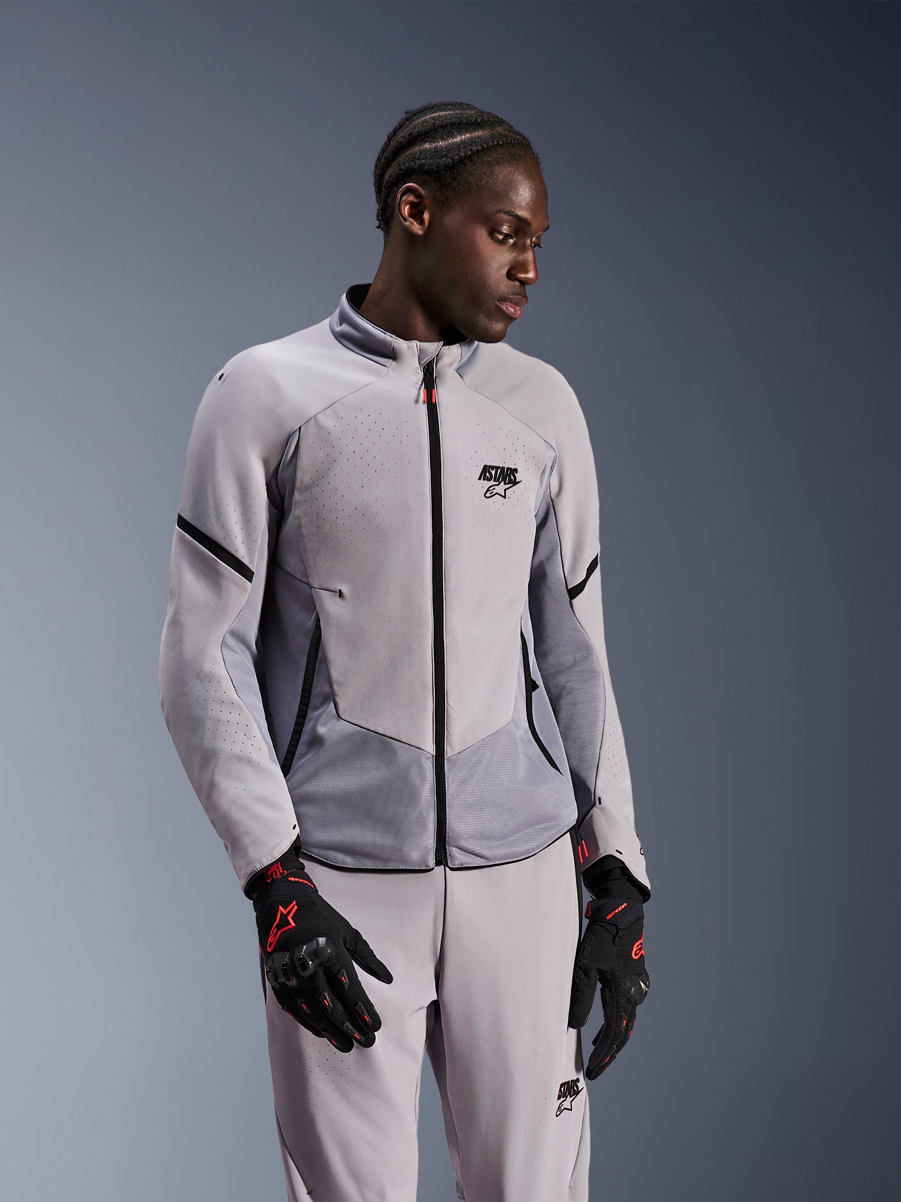 Veste Alpinestars Aeroshell Airflow, gris anthracite moyen, portée par un mannequin avec un pantalon textile assorti et des gants noirs avec des accents rouges, dotée de zones perforées au laser et de panneaux en maille pour la ventilation, design à coupe sportive pour la conduite moto.
