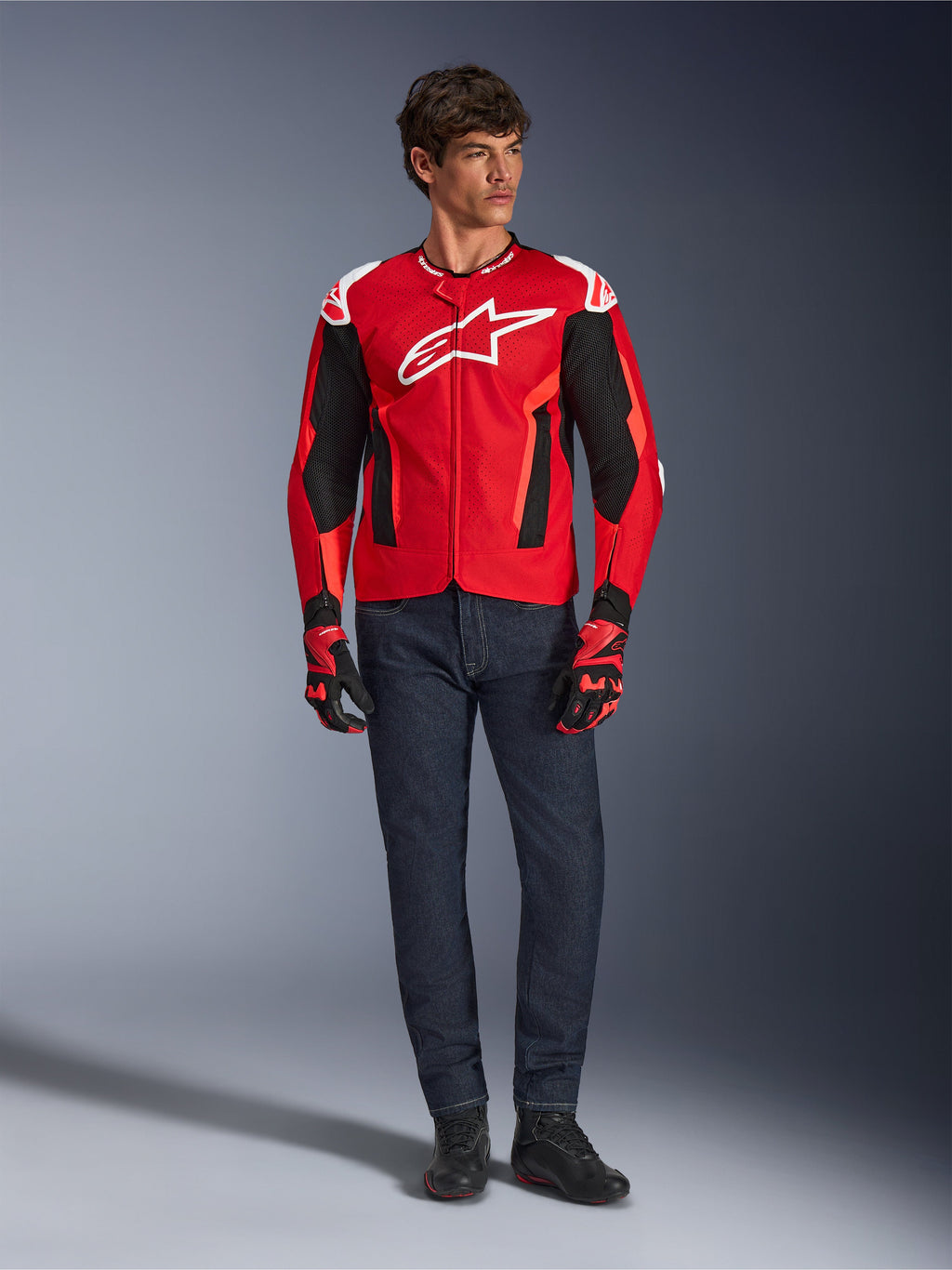 Veste Alpinestars T-GP Air, rouge vif, rouge fluo et noir, modèle masculin portant une tenue de moto complète avec jean bleu foncé et gants assortis, veste textile sportive avec panneaux en maille perforée et logo Astars blanc, photo de studio.