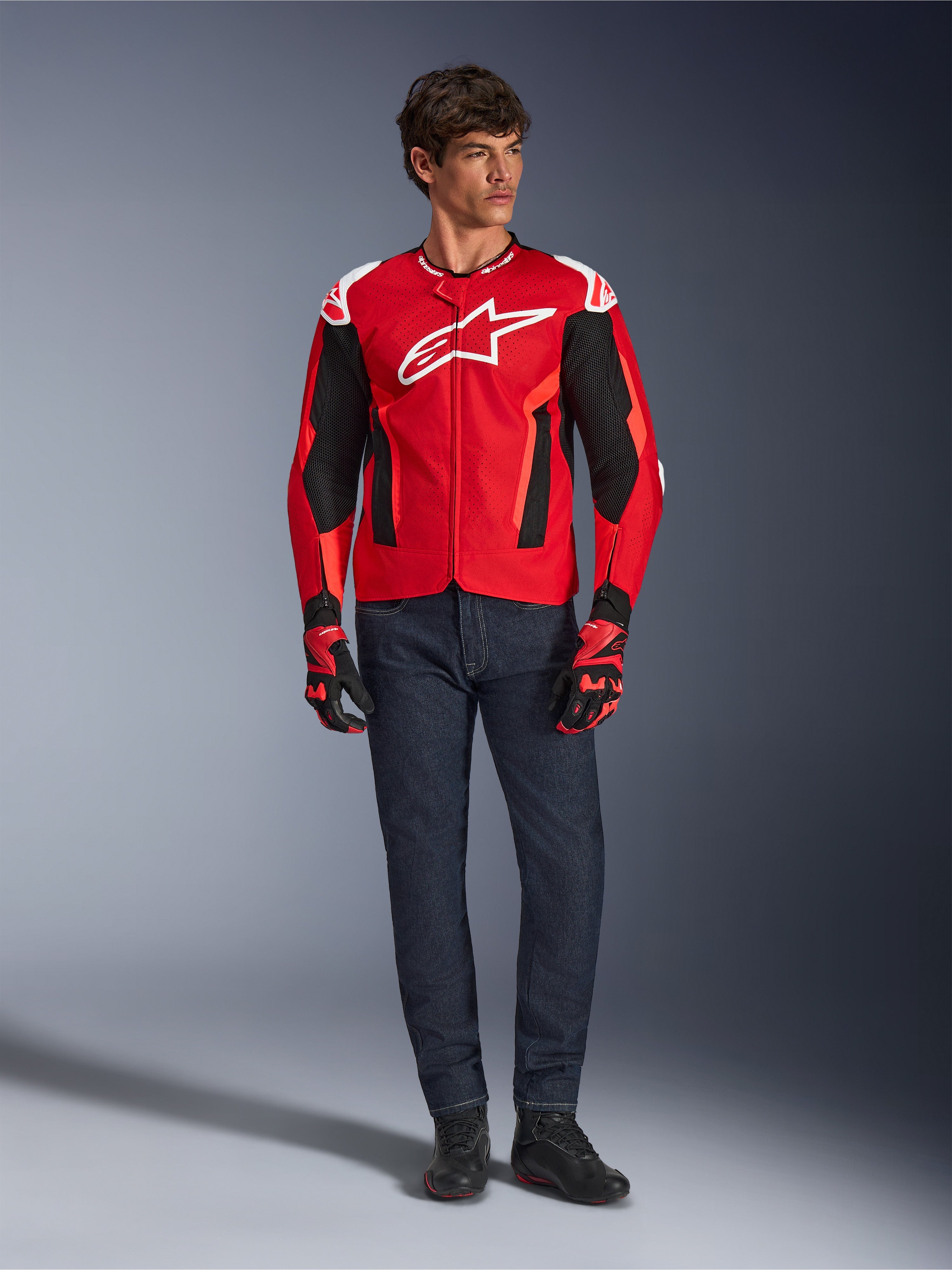 Veste Alpinestars T-GP Air, rouge vif, rouge fluo et noir, modèle masculin portant une tenue de moto complète avec jean bleu foncé et gants assortis, veste textile sportive avec panneaux en maille perforée et logo Astars blanc, photo de studio.