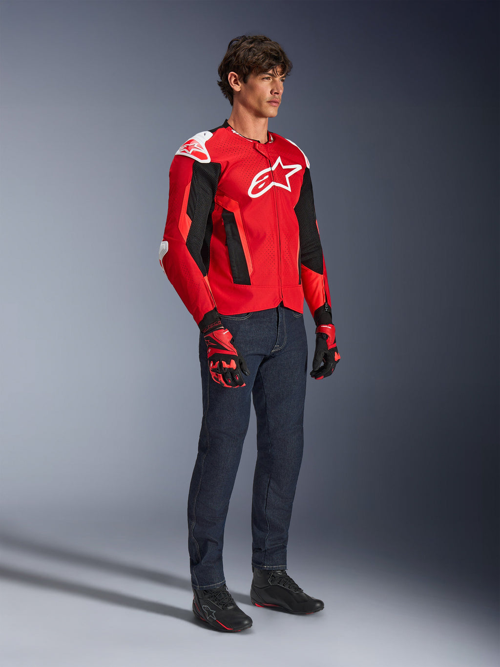 Veste Alpinestars T-GP Air, veste de moto sport, rouge vif et noir avec des accents blancs, portée par un mannequin avec un jean bleu foncé et des gants de course rouges, dotée de panneaux en maille à haut flux et de sliders d'épaule blancs, coupe sport aérodynamique pour la conduite par temps chaud.