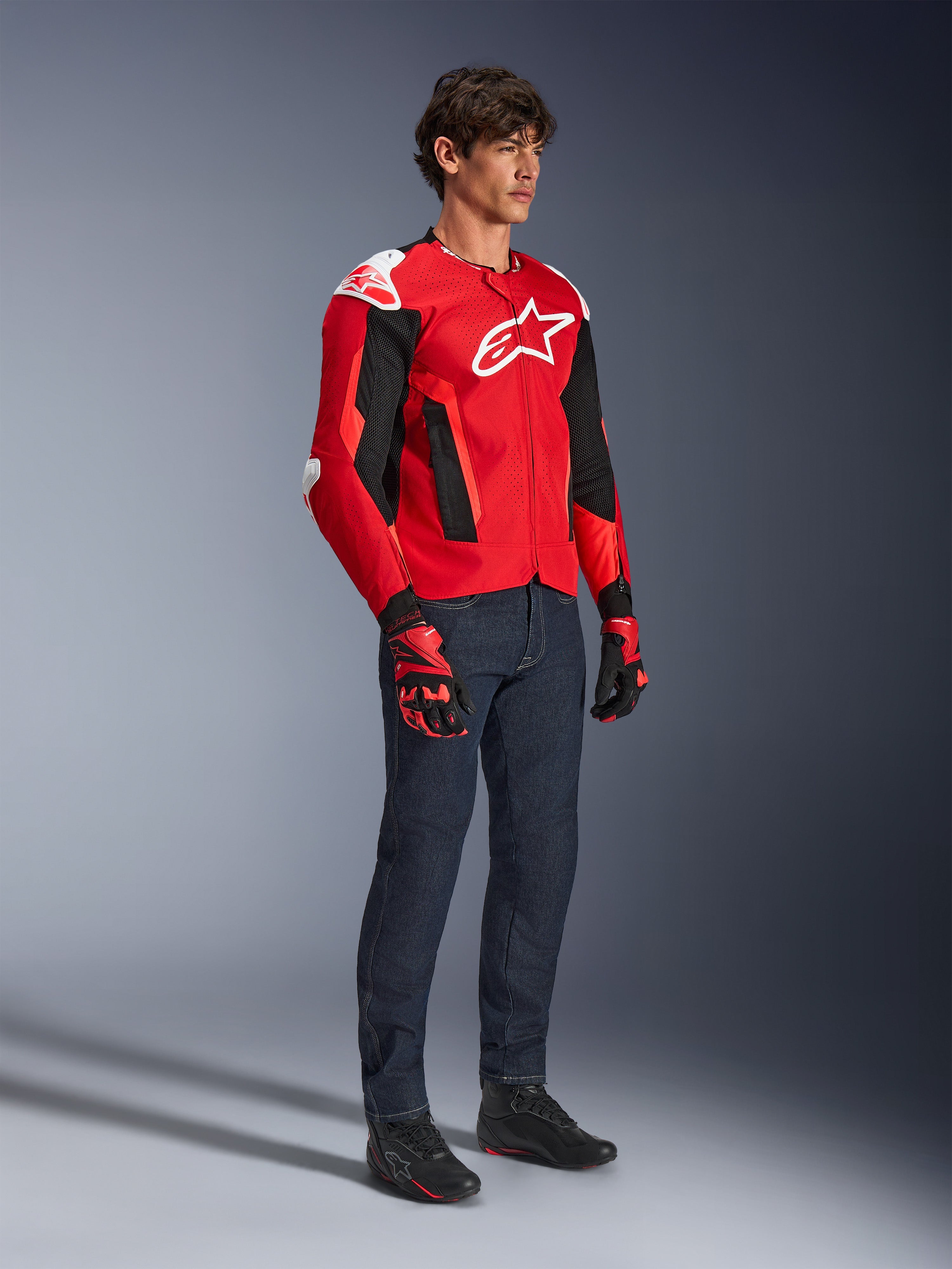 Veste Alpinestars T-GP Air, veste de moto sport, rouge vif et noir avec des accents blancs, portée par un mannequin avec un jean bleu foncé et des gants de course rouges, dotée de panneaux en maille à haut flux et de sliders d'épaule blancs, coupe sport aérodynamique pour la conduite par temps chaud.