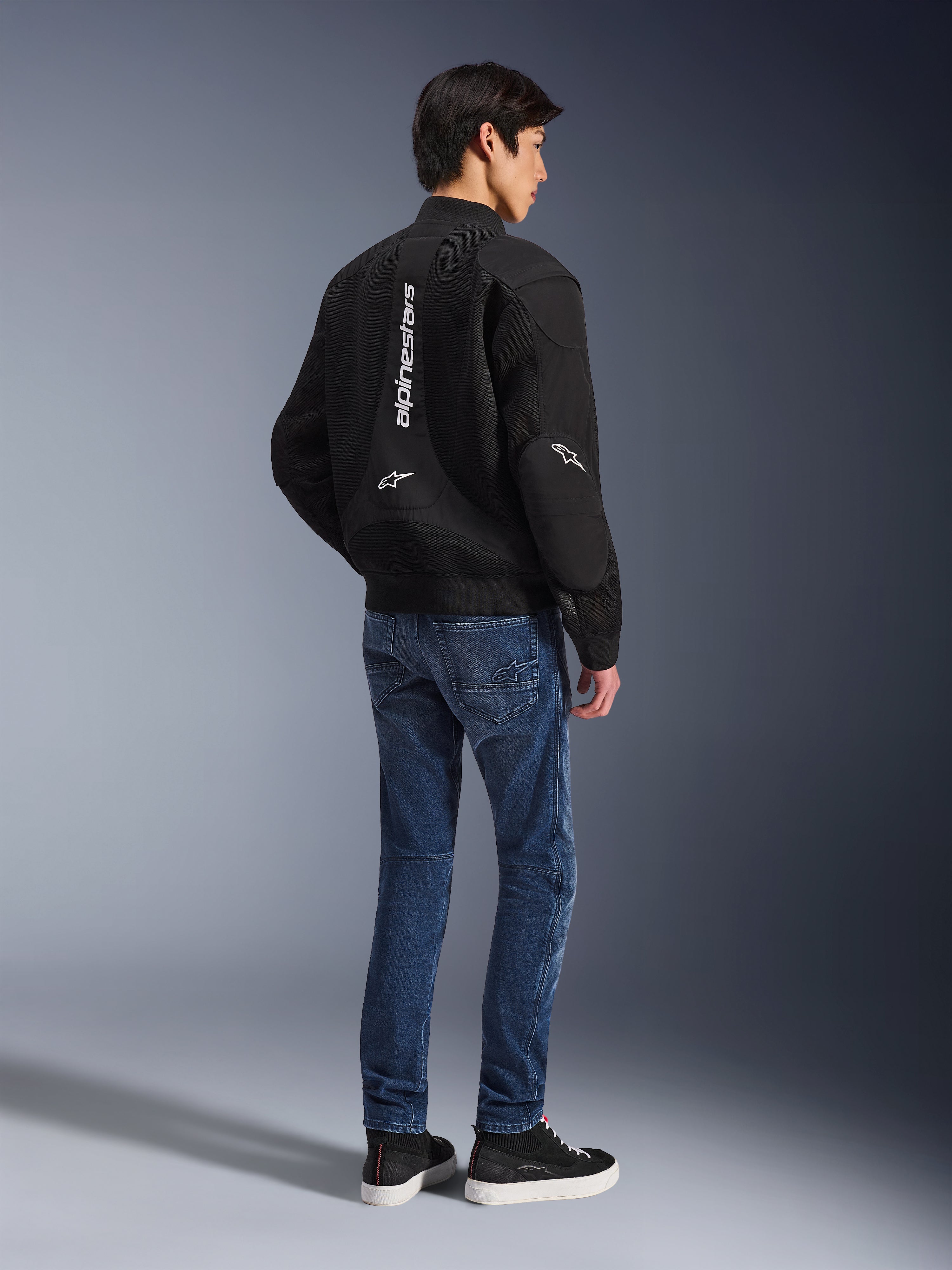 Modèle portant la veste Alpinestars Flight Air noire, vue de dos, associée à un jean de moto bleu et des baskets noires, avec marquage Alpinestars vertical blanc et panneaux de ventilation en maille 3D.