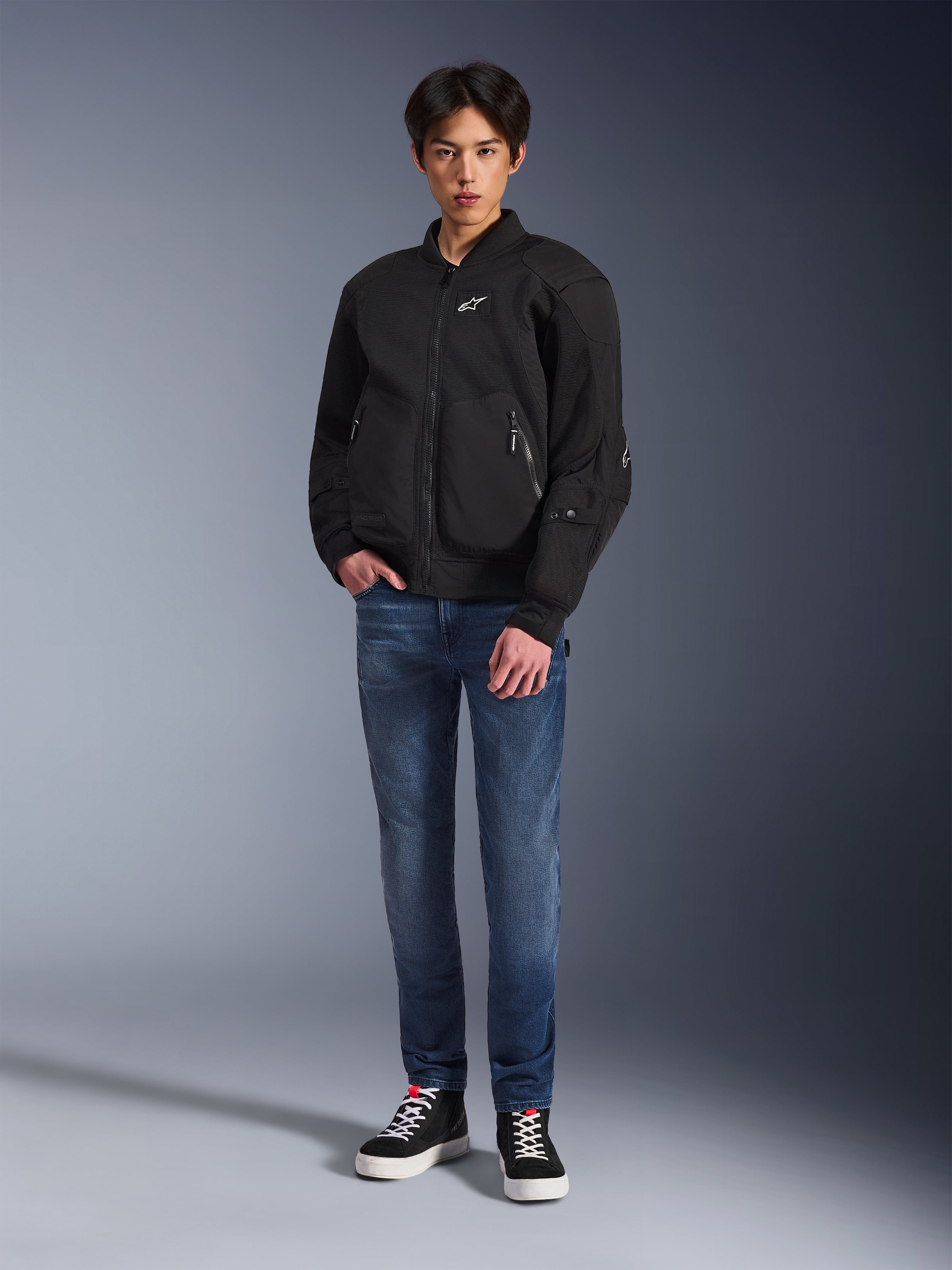 Modèle portant la veste Alpinestars Flight Air en noir, une veste de moto urbaine de style bomber avec des panneaux en maille 3D et une fermeture éclair frontale, associée à un jean bleu et des baskets noires, équipement de moto contemporain d'inspiration streetwear.