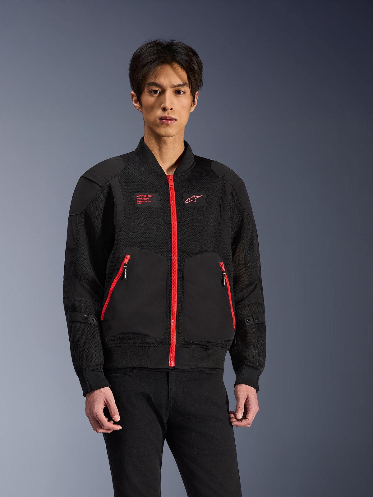 Alpinestars Flight Air Jacket in Black and Bright Red, urban bomber-style motorcycle jacket worn by a model, featuring mesh ventilation panels, a prominent red front zipper, red pocket details, and chest branding, styled with black pants