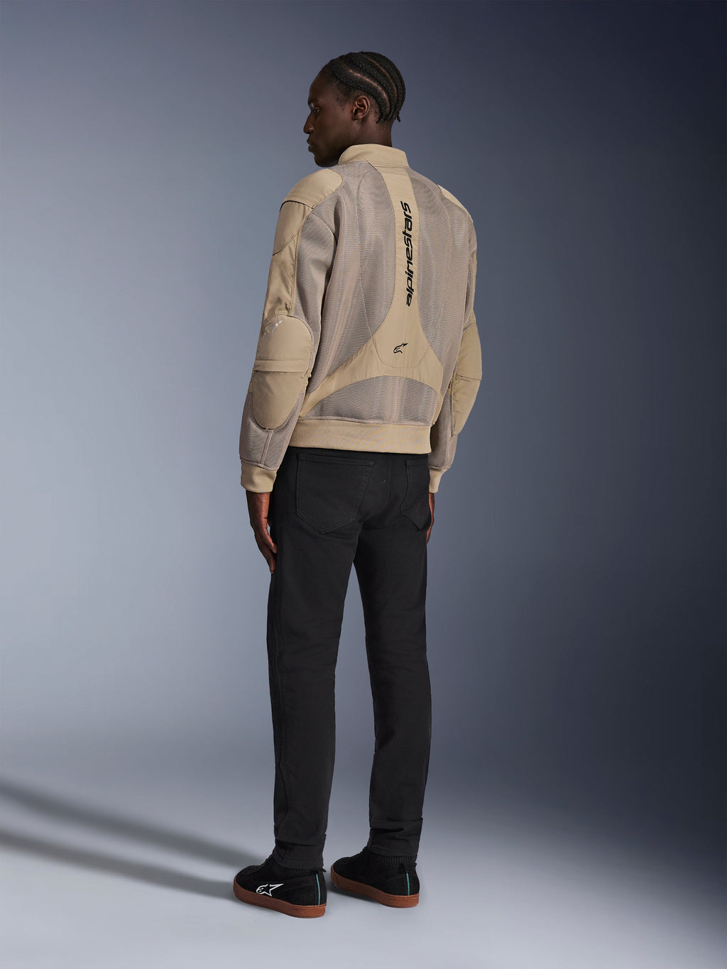 Veste Alpinestars Flight Air, veste de moto urbaine en textile, marron, vue arrière montrant de larges panneaux en maille 3D et un logo vertical, silhouette de style bomber avec col et ourlet côtelés, associée à un pantalon noir et des chaussures noires pour un style de conduite d'inspiration urbaine.
