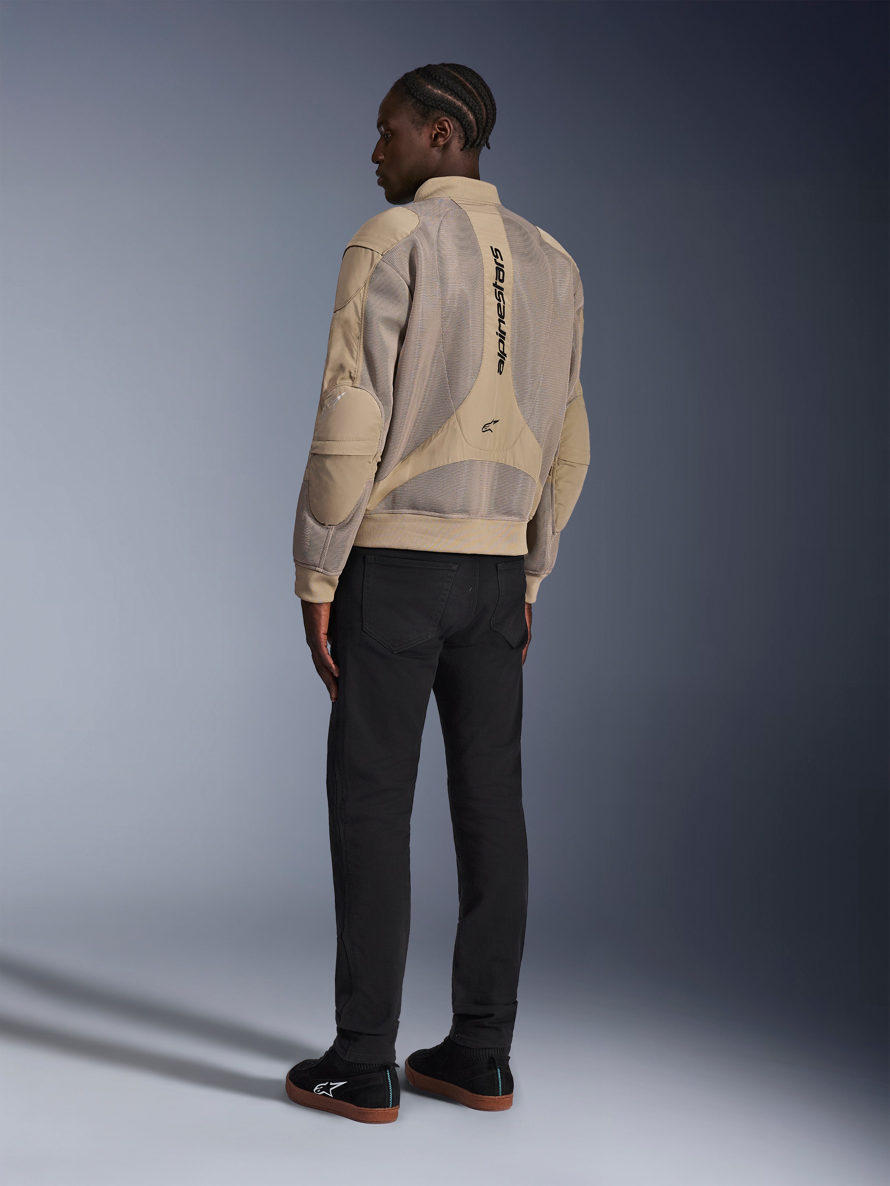 Veste Alpinestars Flight Air, veste de moto urbaine en textile, marron, vue arrière montrant de larges panneaux en maille 3D et un logo vertical, silhouette de style bomber avec col et ourlet côtelés, associée à un pantalon noir et des chaussures noires pour un style de conduite d'inspiration urbaine.