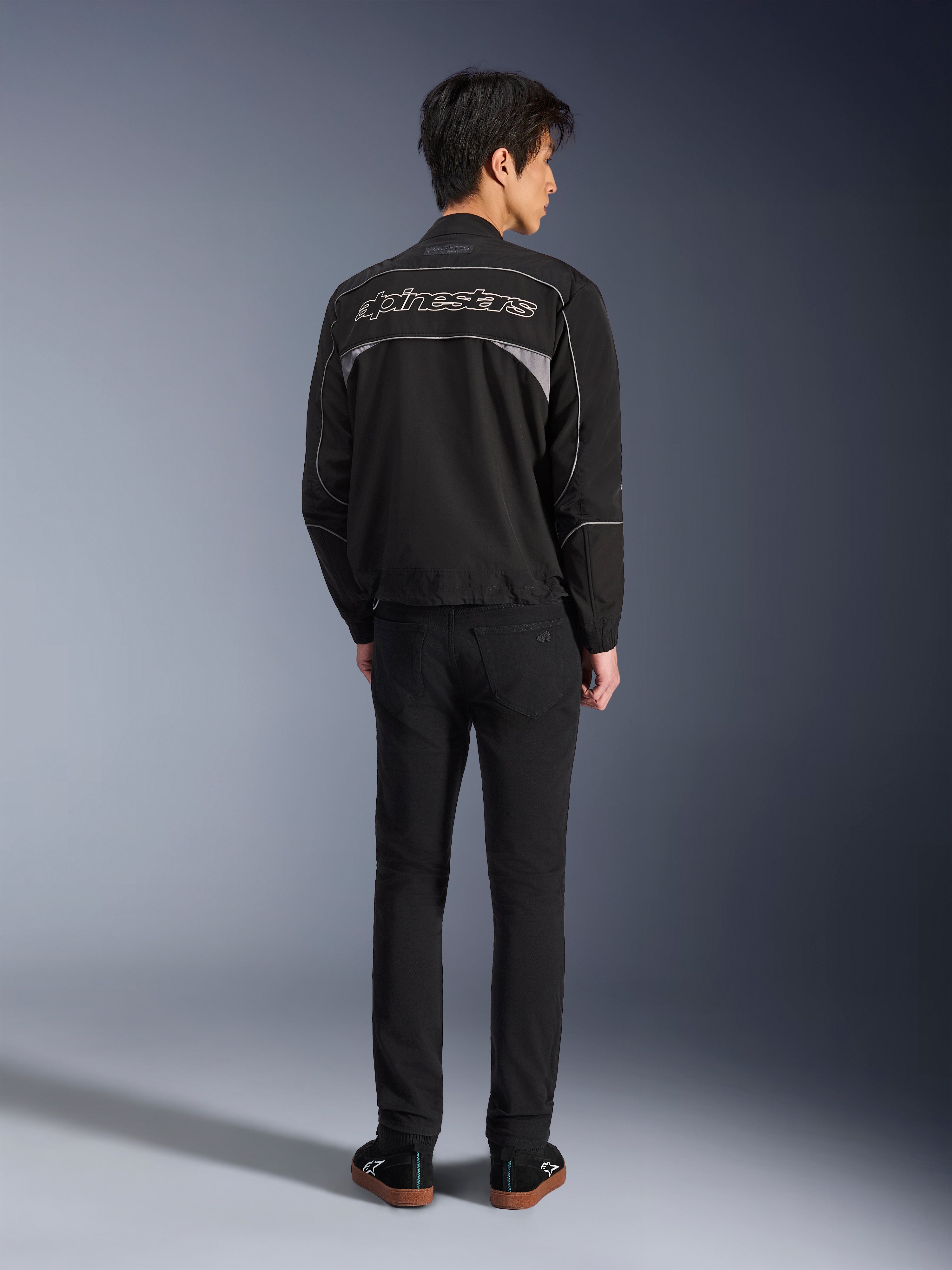 Veste Alpinestars Unite, veste de moto noire et gris foncé, vue arrière montrant le logo Alpinestars sur le dos, panneaux réfléchissants gris, design urbain minimaliste, portée avec un pantalon noir et des chaussures noires