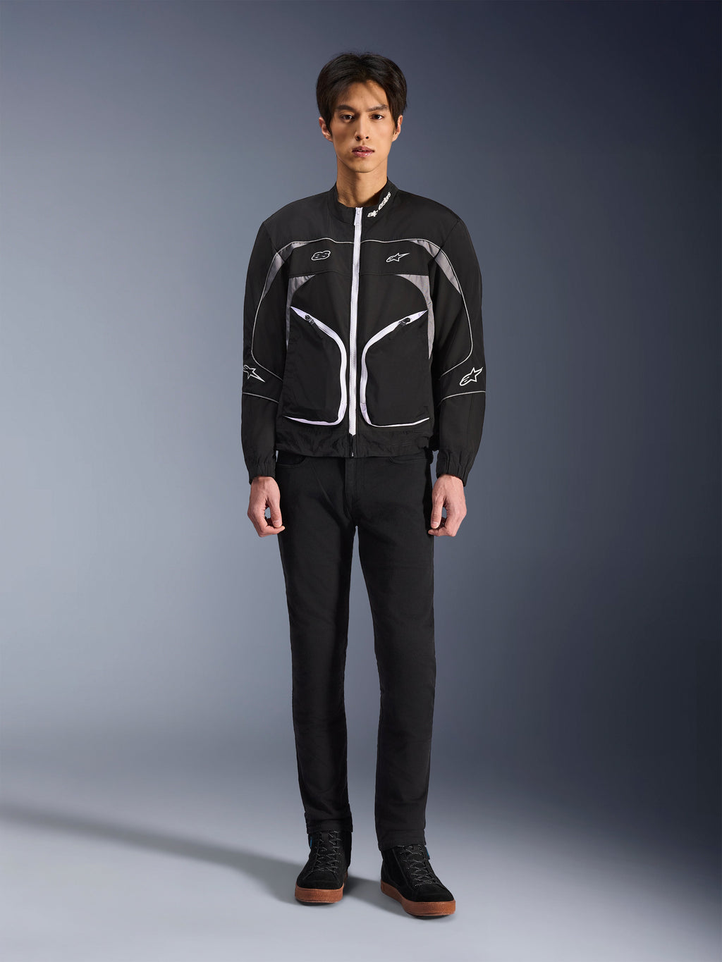 Veste Alpinestars Unite, veste de moto, noir et gris foncé, portée par un mannequin avec un pantalon noir et des bottes noires, dotée de fermetures éclair contrastées blanches, de liserés réfléchissants et d'un design urbain minimaliste