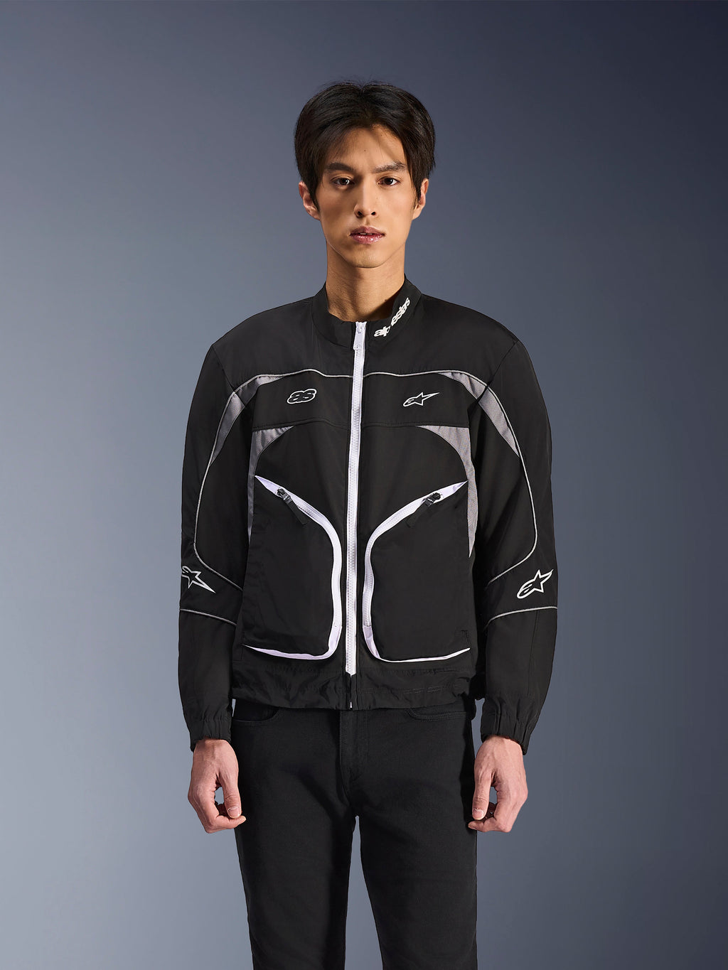 Veste Alpinestars Unite, veste de moto urbaine minimaliste, noir et gris foncé avec des accents blancs, dotée d'une fermeture éclair frontale blanche, de liserés réfléchissants et du logo Alpinestars sur la poitrine et les manches, style streetwear contemporain pour les motards.