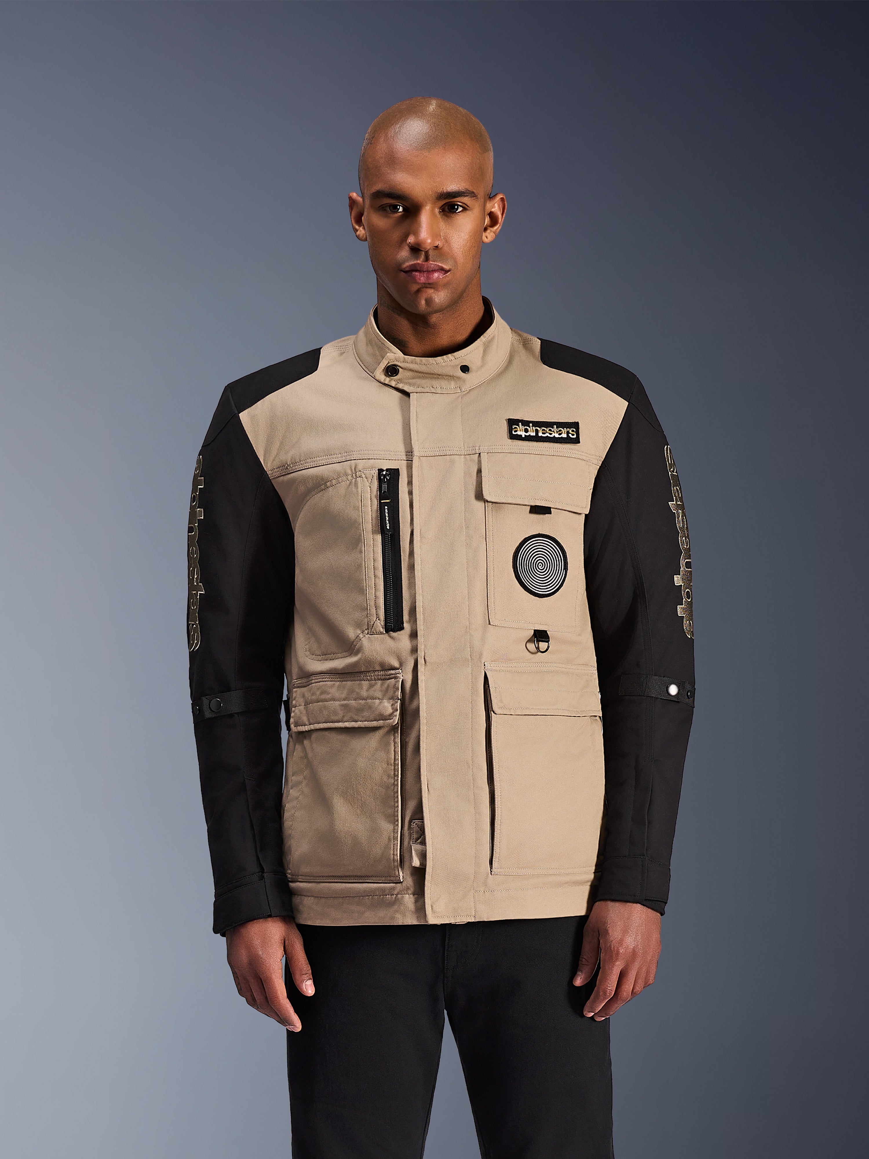Veste Alpinestars Dice, veste de moto urbaine, beige et noire, esthétique workwear robuste avec plusieurs poches utilitaires et des écussons sur la poitrine, col haut avec fermeture à bouton, logos imposants sur les manches, portée par un mannequin homme