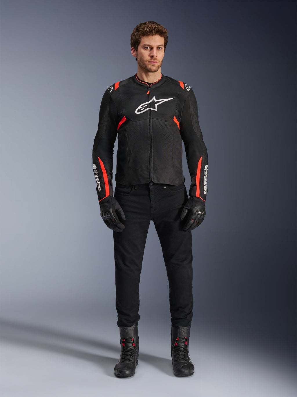 Modèle masculin portant la veste Alpinestars T-SPS Air v2, veste de moto en textile noir avec accents rouge fluo, logo blanc sur la poitrine, portée avec un pantalon, des gants et des bottes noirs, style de conduite urbain sport