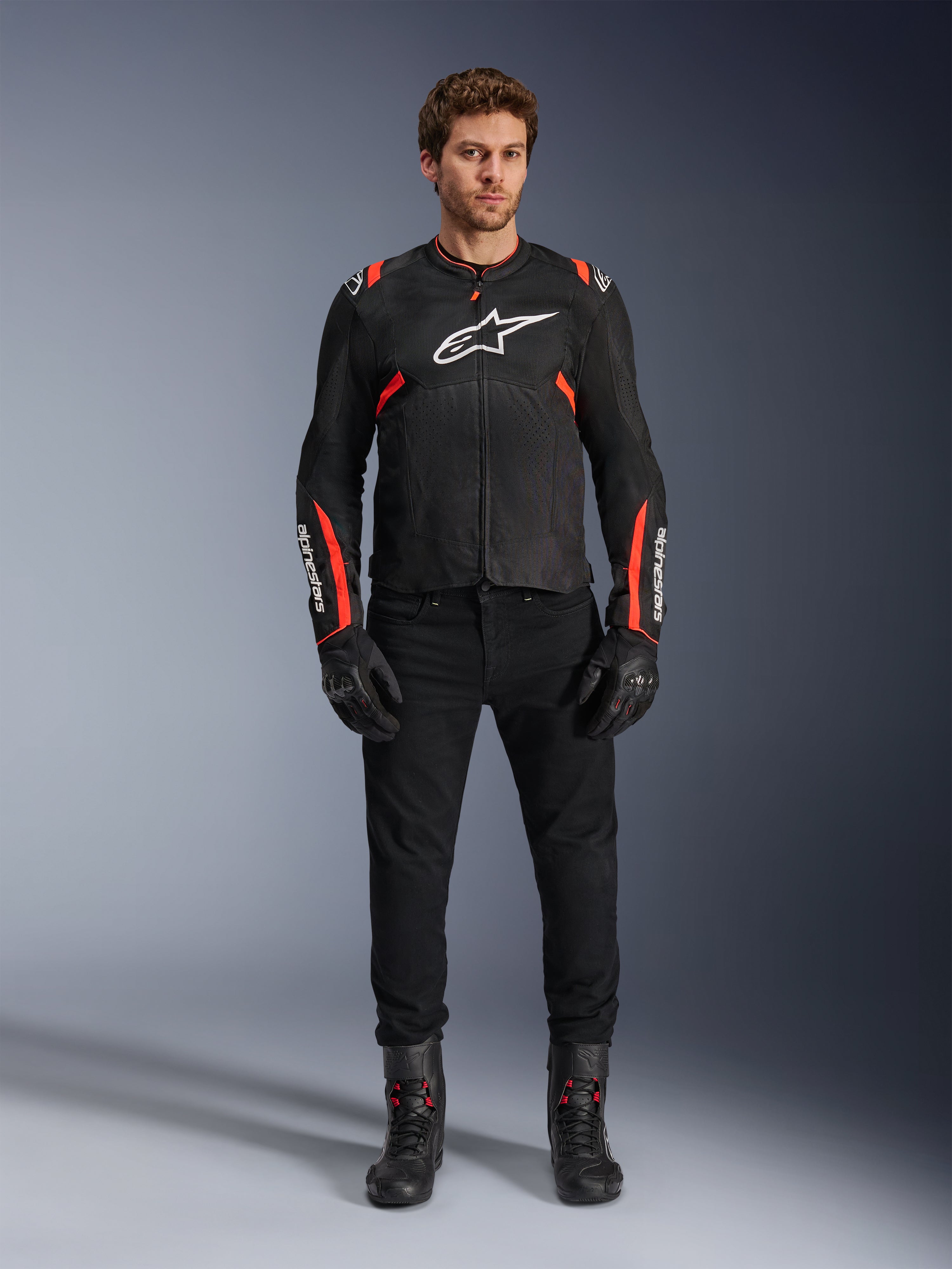 Modèle masculin portant la veste Alpinestars T-SPS Air v2, veste de moto en textile noir avec accents rouge fluo, logo blanc sur la poitrine, portée avec un pantalon, des gants et des bottes noirs, style de conduite urbain sport