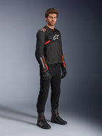 Modèle masculin portant la veste Alpinestars T-Sps Air V2, noir avec accents rouge fluo, associée à un pantalon de moto noir, des gants noirs et des bottes noires, mettant en valeur un design sportif avec des panneaux en mesh et des logos Alpinestars blancs.