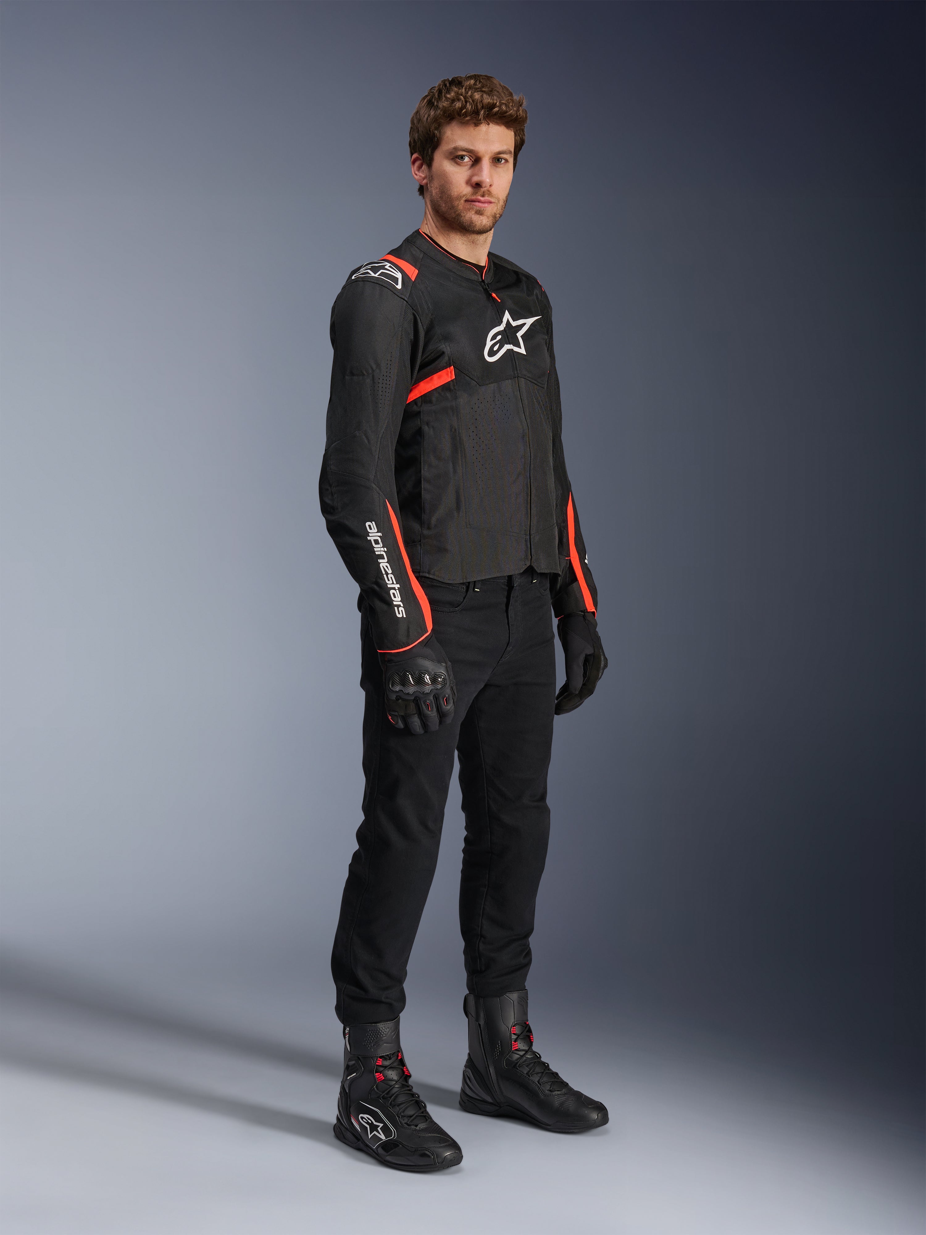 Modèle masculin portant la veste Alpinestars T-Sps Air V2, noir avec accents rouge fluo, associée à un pantalon de moto noir, des gants noirs et des bottes noires, mettant en valeur un design sportif avec des panneaux en mesh et des logos Alpinestars blancs.