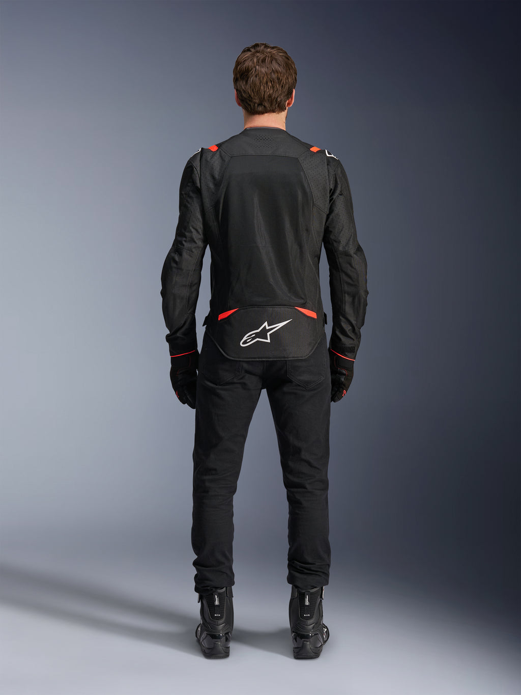 Veste Alpinestars T-SPS Air v2 noir et rouge fluo, vue arrière montrant de grands panneaux de ventilation en mesh et le logo Alpinestars blanc dans le bas du dos, portée avec un pantalon de moto et des bottes noirs.