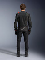 Veste Alpinestars T-SPS Air v2 noir et rouge fluo, vue arrière montrant de grands panneaux de ventilation en mesh et le logo Alpinestars blanc dans le bas du dos, portée avec un pantalon de moto et des bottes noirs.
