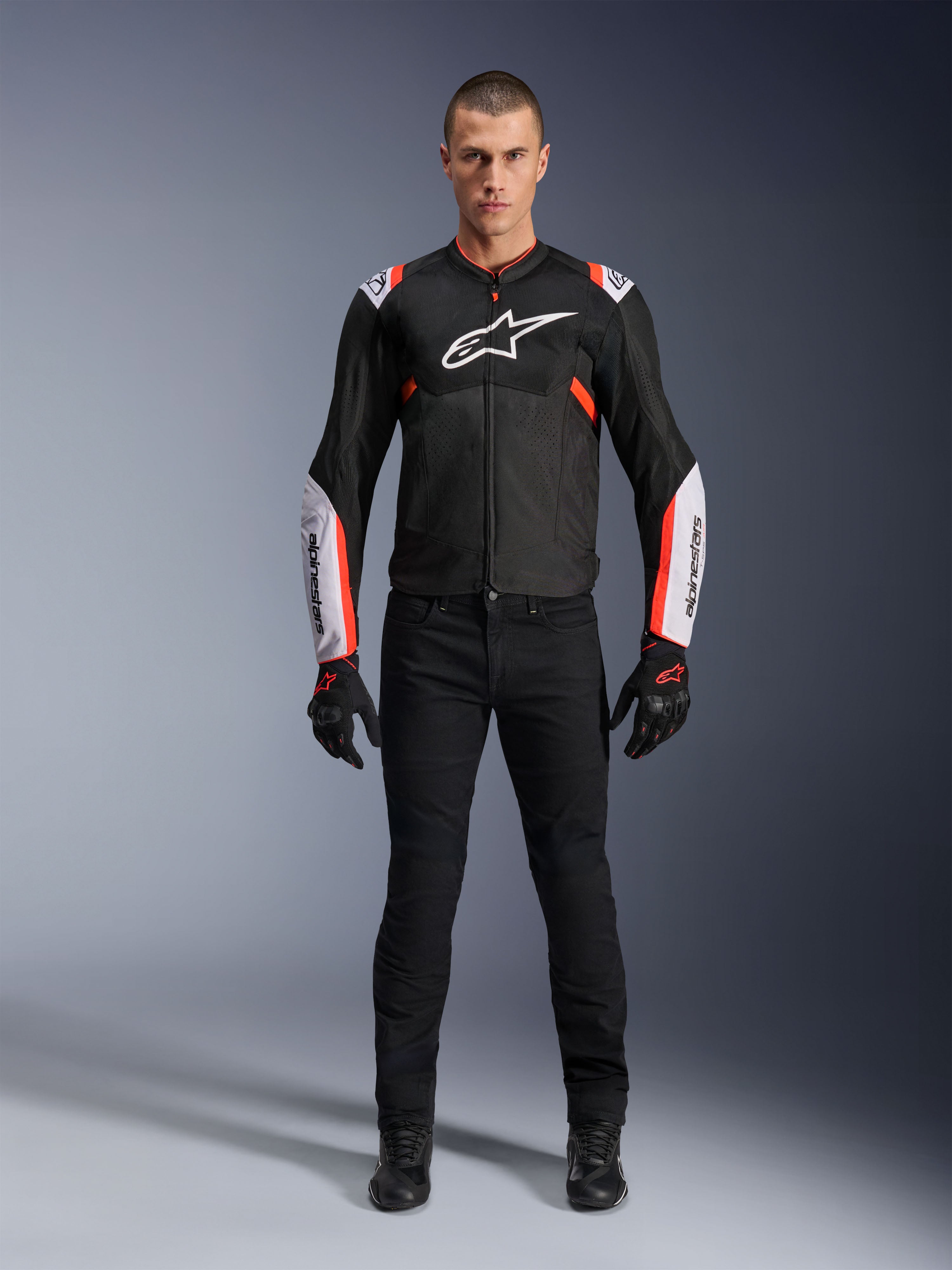Modèle masculin portant la veste Alpinestars T-Sps Air V2 en Noir Blanc Rouge Fluo, dotée de larges panneaux en mesh technique et d'un logo blanc sur la poitrine et les manches, associée à un pantalon de moto technique et des gants noirs, équipement de moto sport urbain