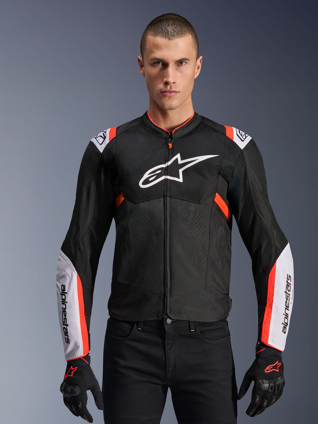 Veste Alpinestars T-Sps Air V2, noire avec des accents blancs et rouge fluo, veste de moto sportive, homme portant un modèle ventilé en mesh avec un grand logo sur la poitrine et des manches marquées, associée à des gants et un pantalon noirs