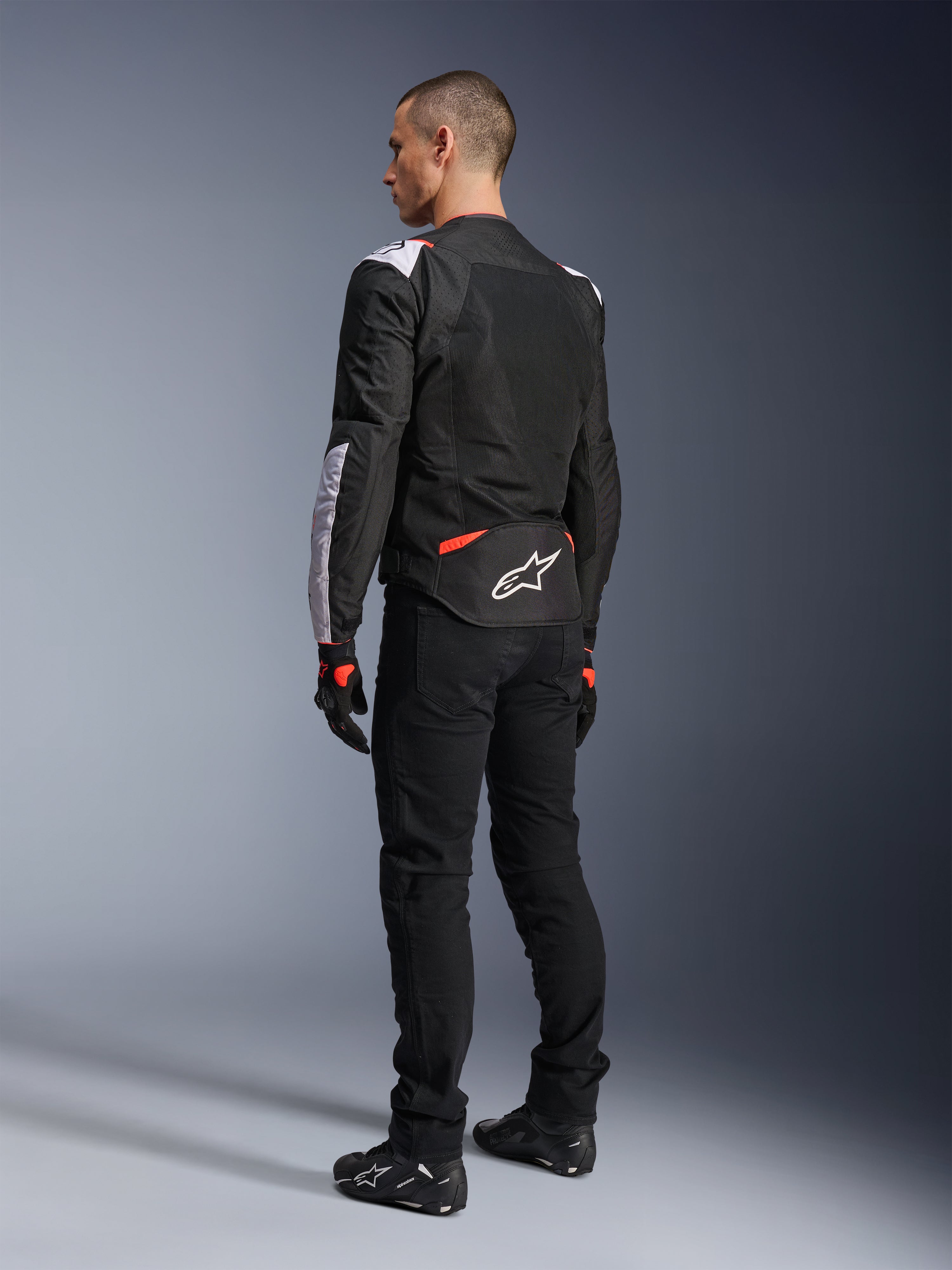 Veste Alpinestars T-Sps Air V2, veste de moto textile, noir, blanc et rouge fluo, vue arrière sur modèle, empiècements en mesh pour la ventilation, logo Astars blanc dans le bas du dos, portée avec un pantalon de moto noir et des gants
