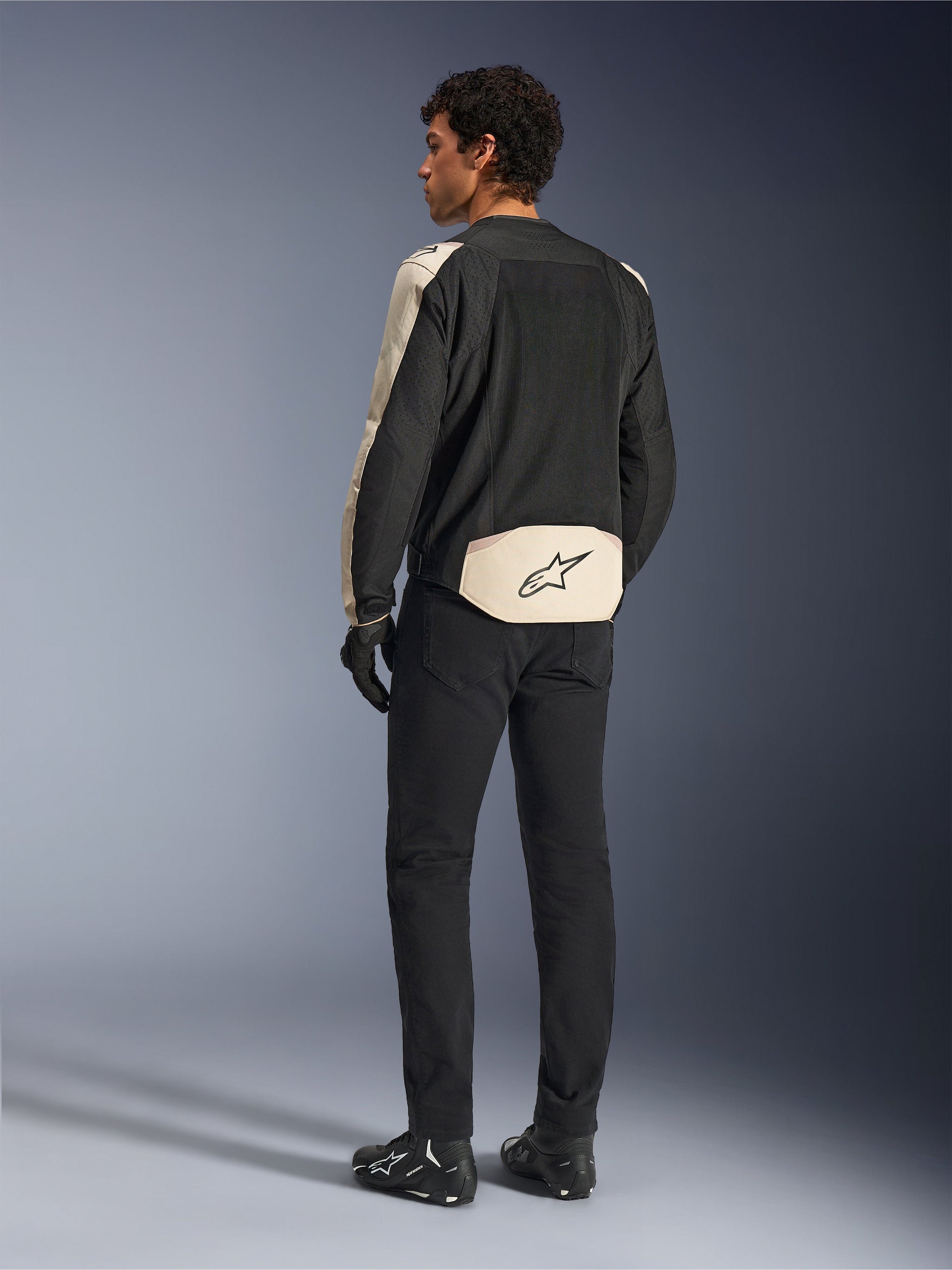 Veste Alpinestars T-SPS Air V2, veste de moto en textile, Noir Mastic Aluminium, vue de dos portée par un modèle, dotée de larges panneaux en maille technique pour la circulation de l'air, accents beige clair sur les manches et le bas du dos avec le logo Alpinestars, associée à un pantalon et des bottes noirs.