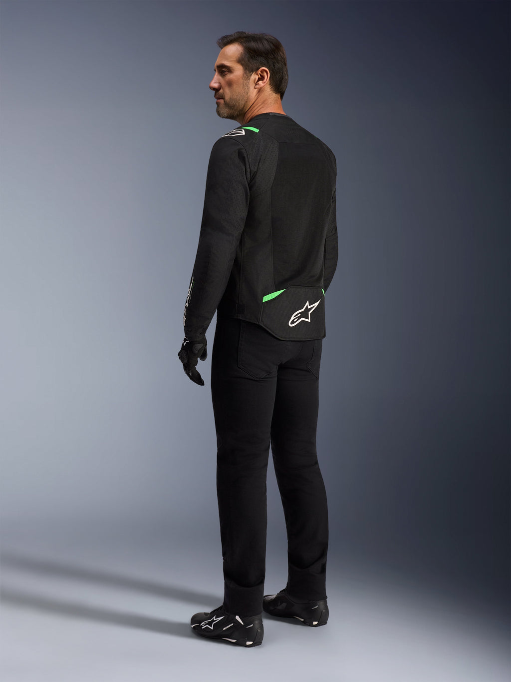 Un homme portant la veste Alpinestars T-SPS Air V2 en Noir Vert Fluo, présenté de dos et de profil, avec un pantalon de moto noir et des chaussures de moto. La veste présente des panneaux en maille technique ventilée, des accents vert vif sur les épaules et le bas du dos, ainsi qu'un logo étoile blanc à l'arrière.