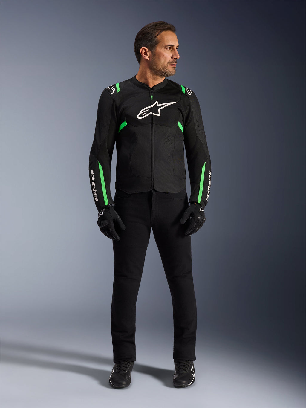 Homme portant la veste Alpinestars T-SPS Air V2 en Noir Vert Fluo, associée à un pantalon noir, des gants et des chaussures de moto, dotée d'une coupe sportive avec des logos blancs et des panneaux en maille pour un look urbain moderne.