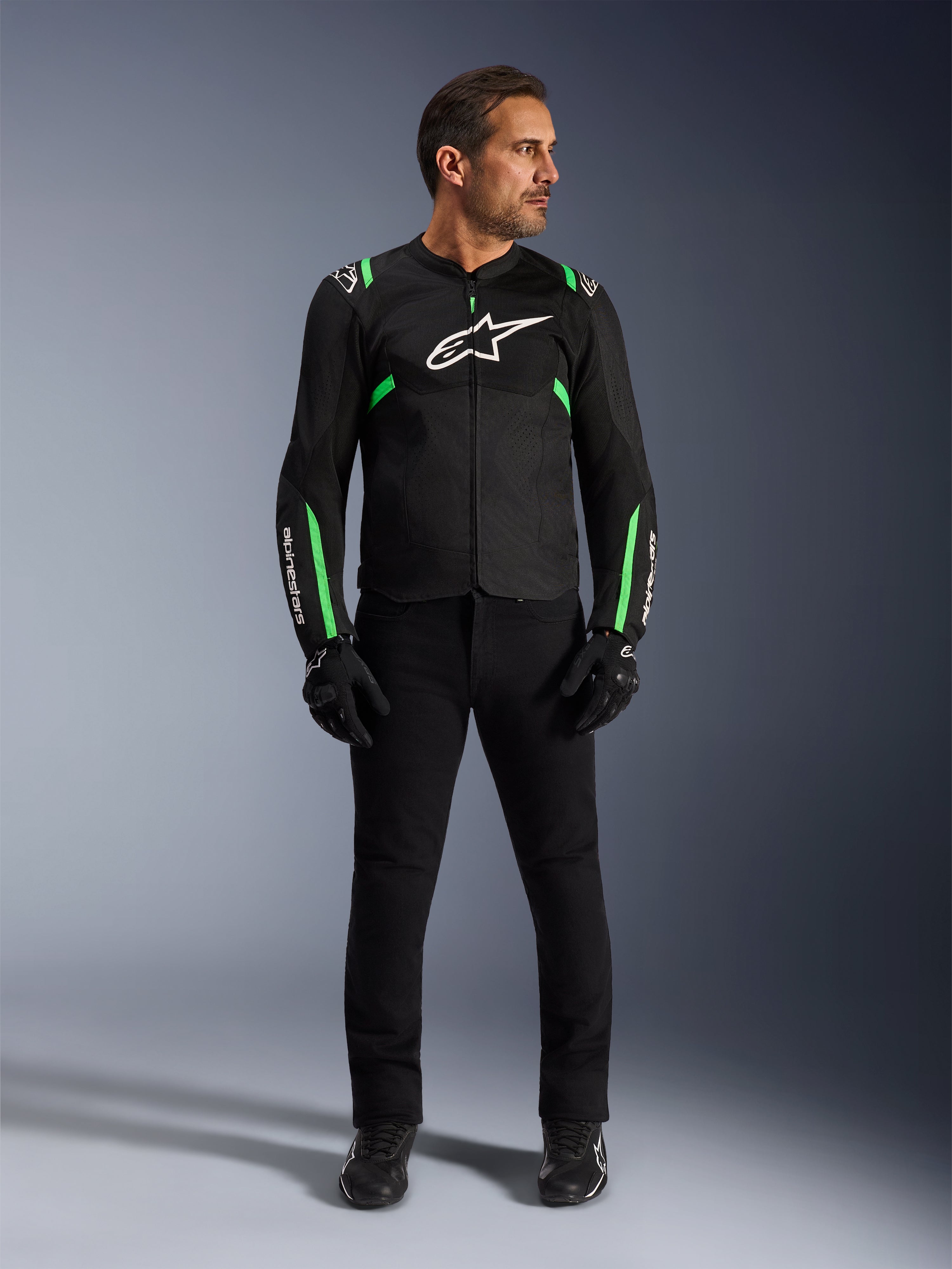Homme portant la veste Alpinestars T-SPS Air V2 en Noir Vert Fluo, associée à un pantalon noir, des gants et des chaussures de moto, dotée d'une coupe sportive avec des logos blancs et des panneaux en maille pour un look urbain moderne.
