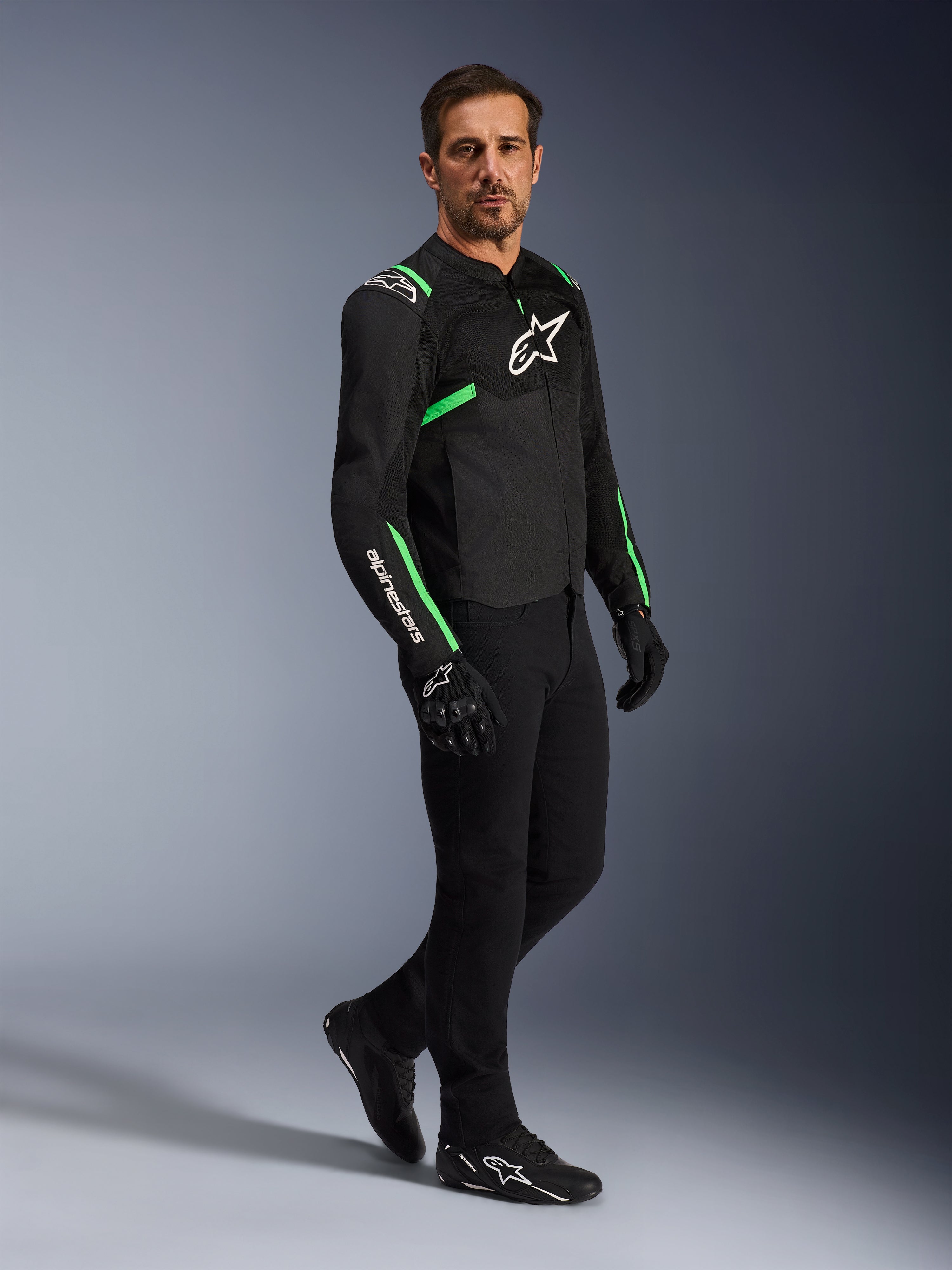 Modèle masculin portant une veste Alpinestars T-SPS Air V2 en noir et vert fluo, associée à un pantalon de moto et des gants noirs, veste textile coupe sport avec panneaux en maille et accents néon, tenue de moto urbaine complète