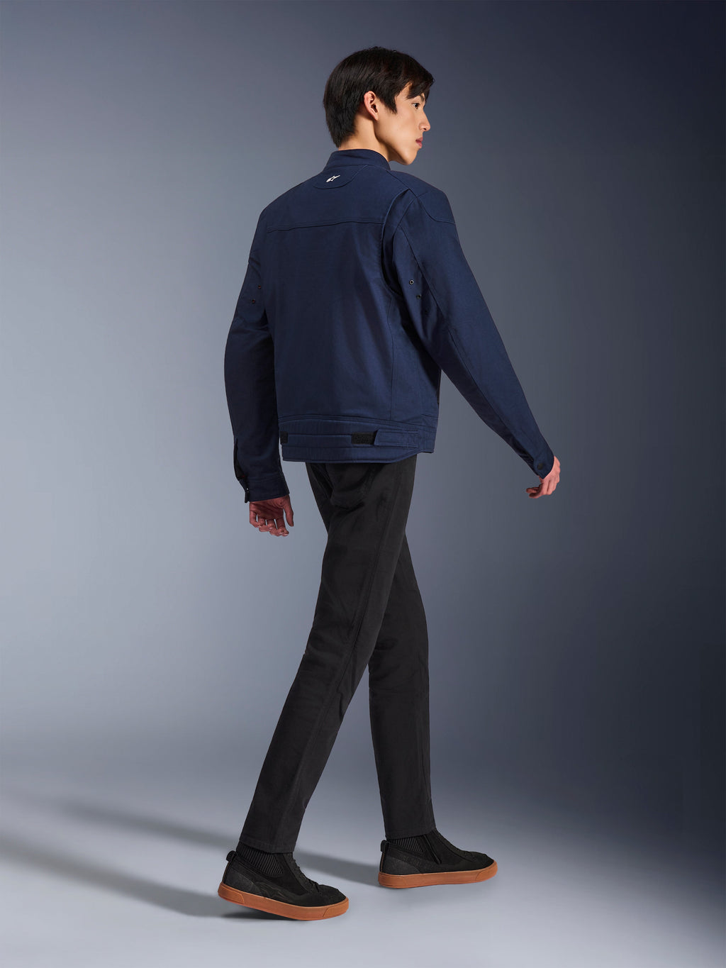 Alpinestars Bruiser Jacket en Bleu Foncé, veste de moto urbaine en textile, vue arrière d'un mannequin qui marche, associée à un pantalon noir et des baskets à semelles en gomme, dotée d'un design inspiré du style workwear et de pattes de réglage à la taille.