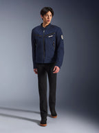 Modèle masculin portant la veste Alpinestars Bruiser en bleu foncé, veste de moto urbaine en textile, associée à un pantalon noir et des baskets noires, esthétique inspirée du vêtement de travail avec poches zippées et écusson circulaire sur la manche, streetwear contemporain pour la conduite à moto.