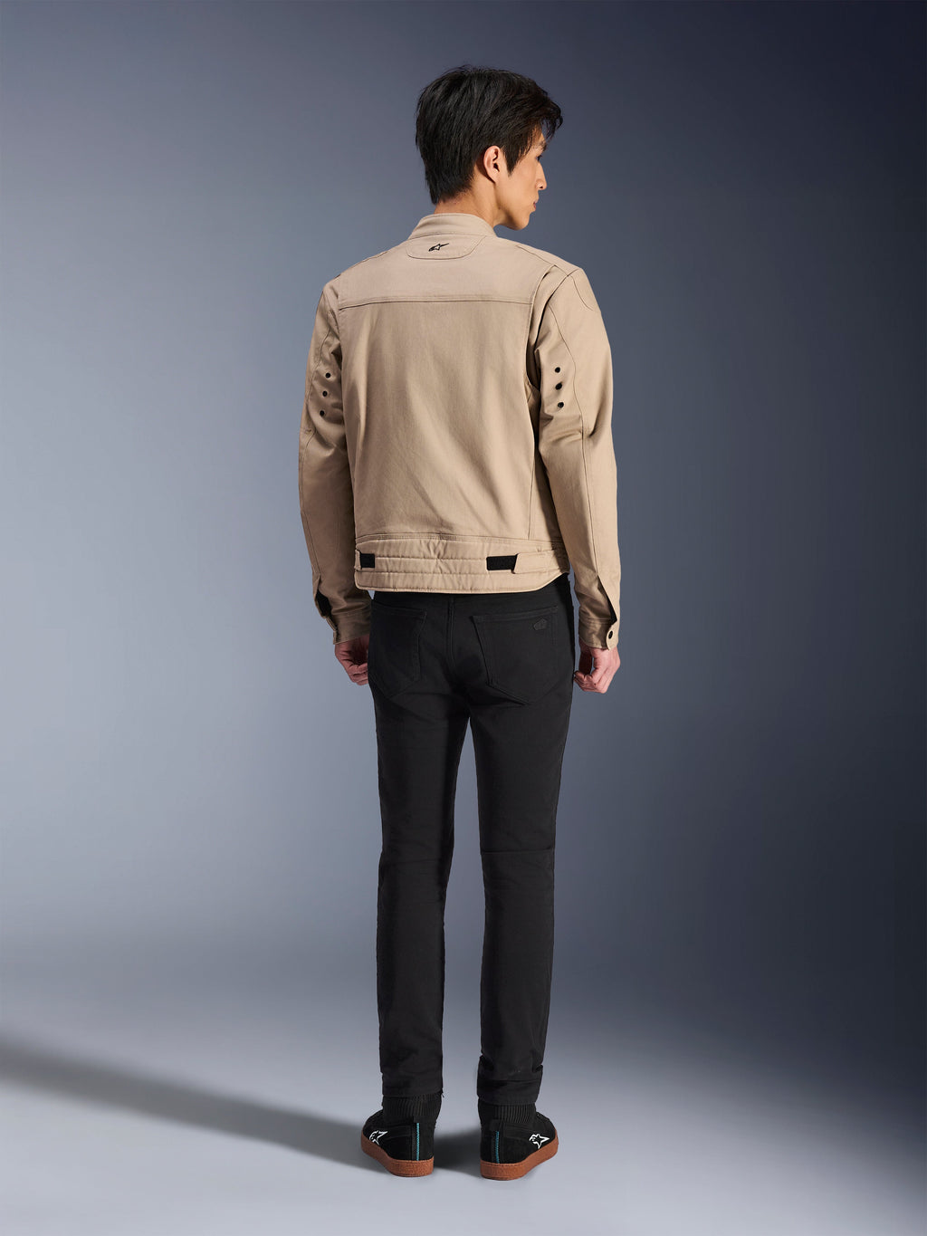 Vue arrière d'un mannequin portant la veste Alpinestars Bruiser marron, veste de moto urbaine au design inspiré du vêtement de travail, avec des œillets métalliques au dos et des ajusteurs à la taille, associée à un pantalon noir et des baskets, style urbain contemporain.