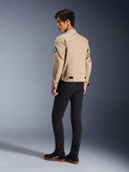 Modèle masculin de dos portant la veste Alpinestars Bruiser en beige, veste de moto urbaine contemporaine au style workwear, avec un écusson circulaire sur la manche, des sangles de réglage à la taille et des épaules plissées, associée à un pantalon noir et des baskets noires.