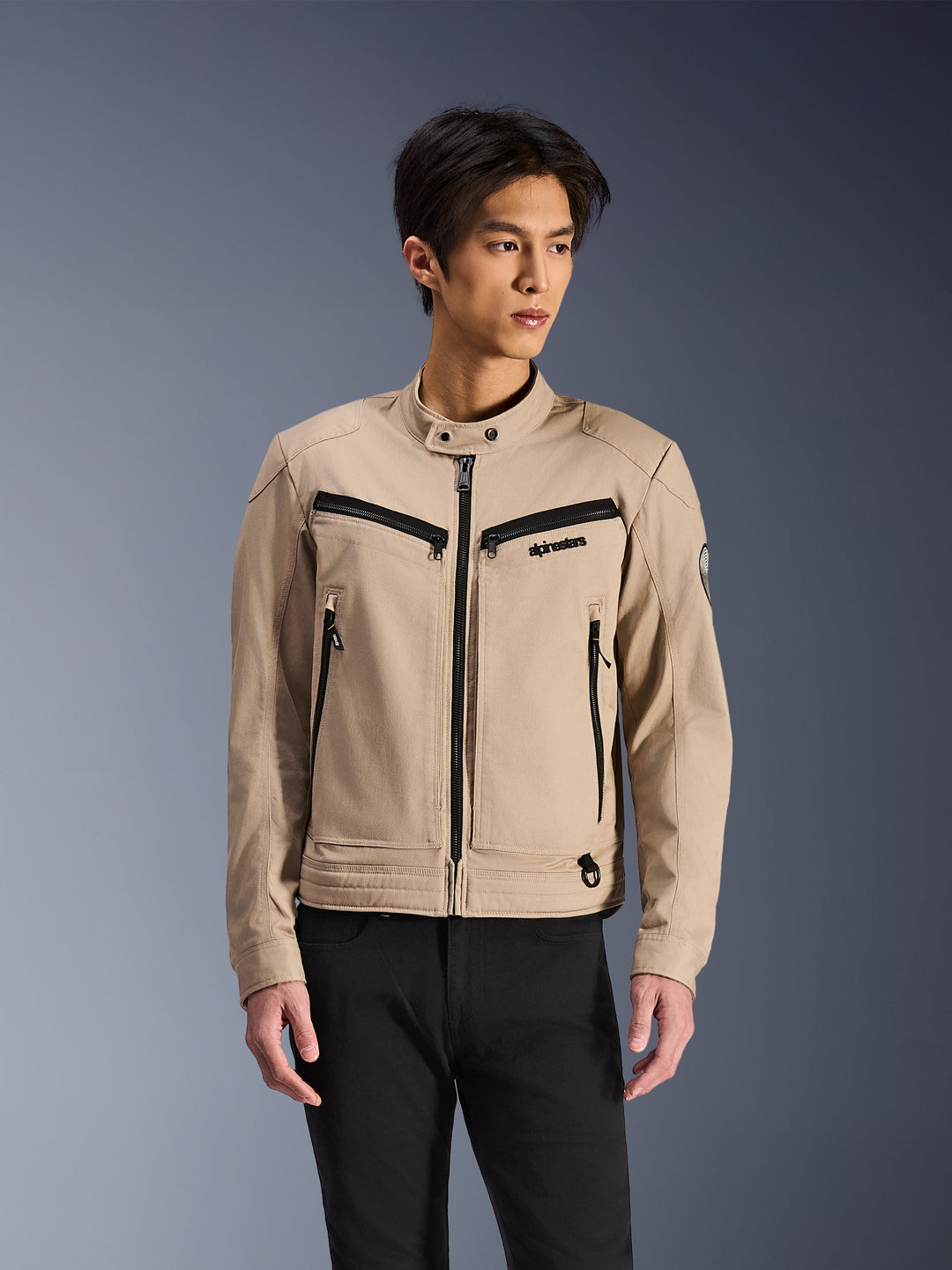 Alpinestars Bruiser Jacket, veste de moto beige portée par un mannequin homme, avec détails de fermetures éclair noires sur la poitrine et les poches latérales, col montant à bouton-pression, vêtement de moto urbain de style workwear, associée à un pantalon noir