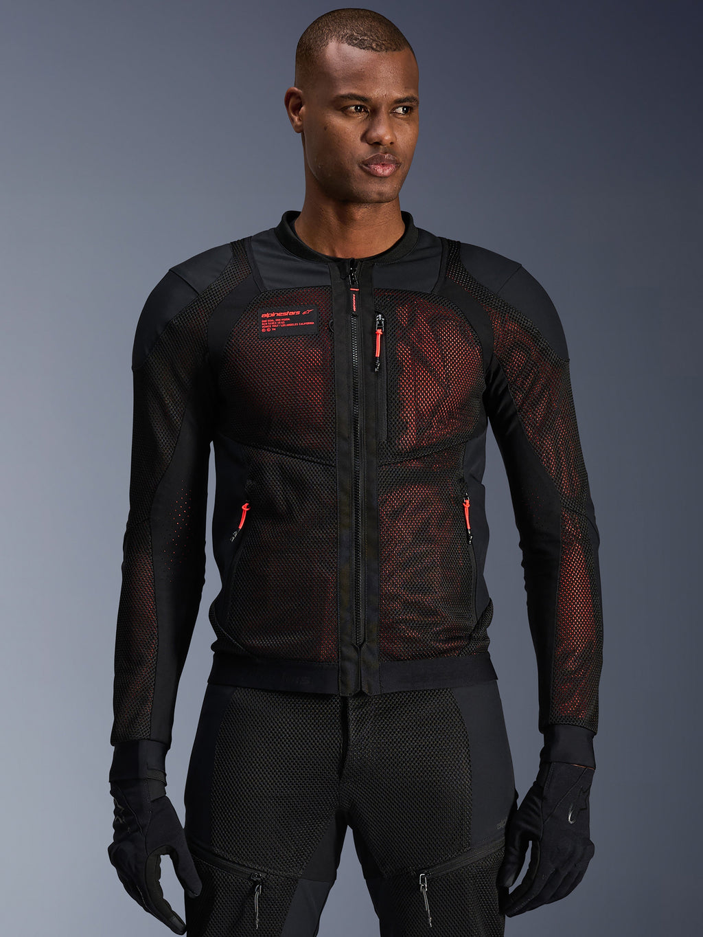 Veste Alpinestars Troop-Air Pro, veste de moto technique en mesh blindée en Noir et Rouge Fluo, portée par un mannequin, dotée de larges panneaux de ventilation en maille, d'une sous-couche rouge et de tirettes rouges, style de conduite urbain.