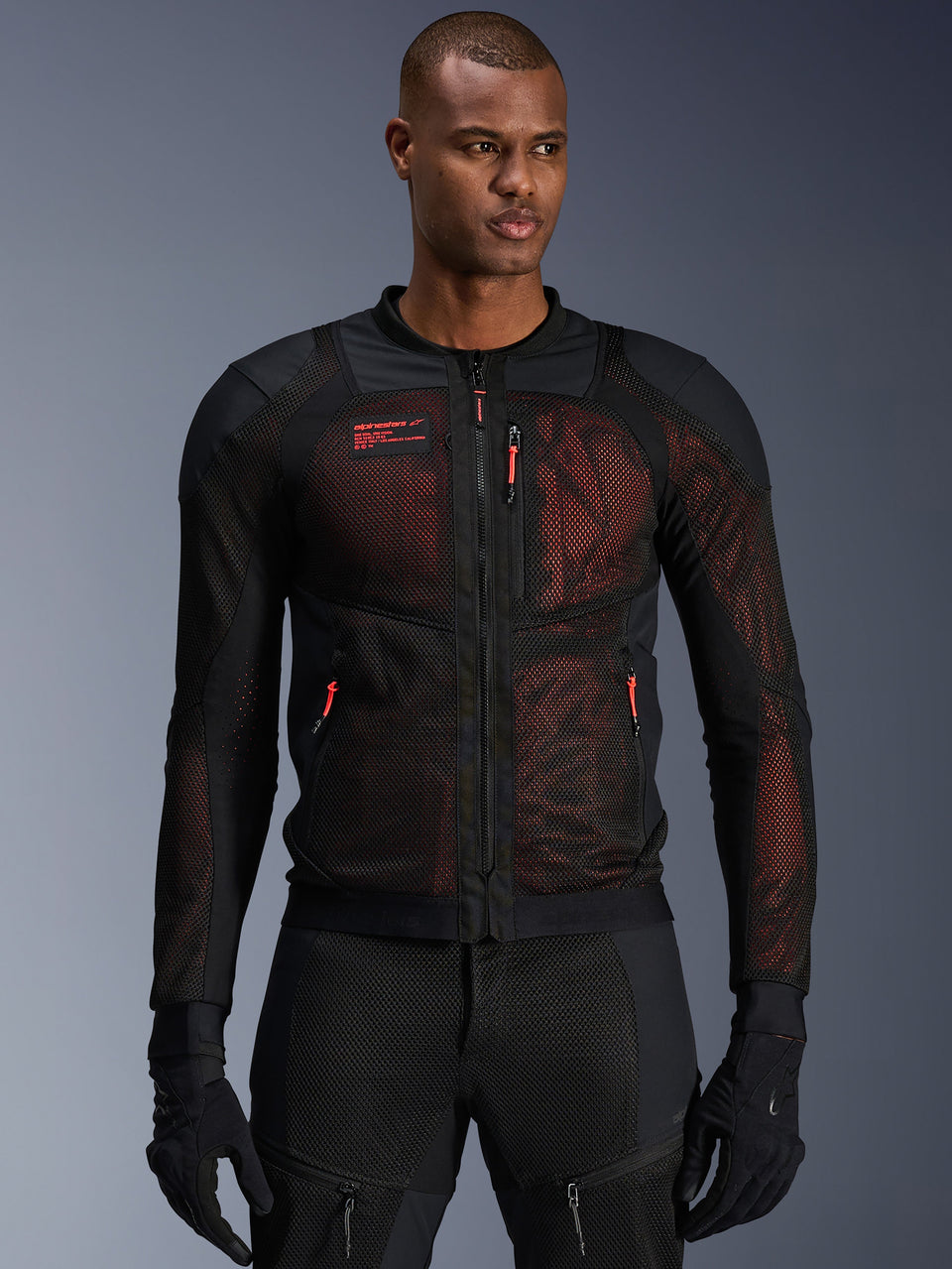 Alpinestars Troop-Air Pro Jacket, technical armored mesh motorcycle jacket in Black and Red Fluo, worn by a model, featuring large mesh ventilation panels, red underlay, and red zipper pulls, urban riding style.