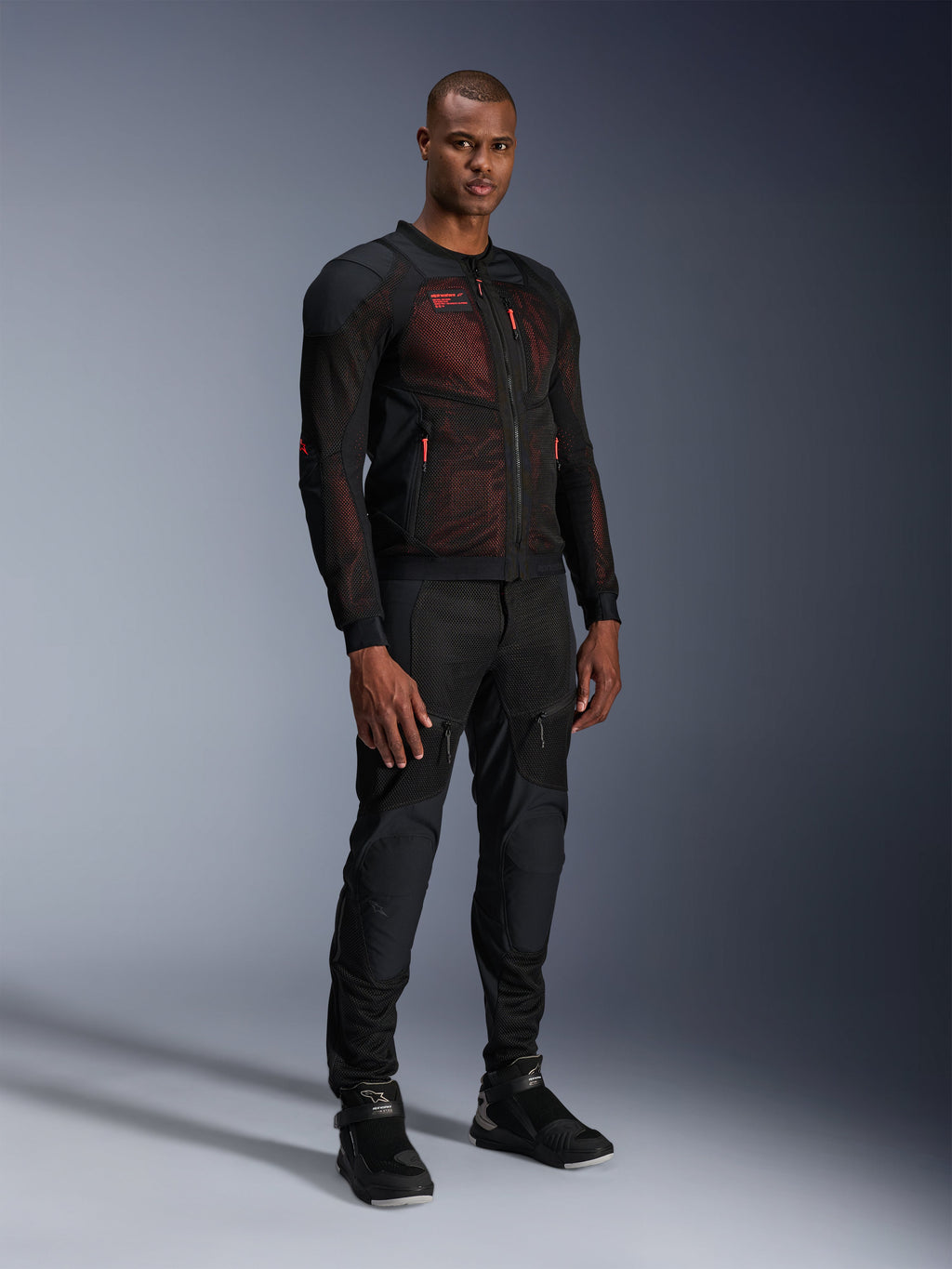 Veste Alpinestars Troop-Air Pro en Noir Rouge Fluo, veste de moto technique en mesh blindée portée par un mannequin homme avec un pantalon blindé noir assorti et des bottes montantes, portrait de studio en pied.