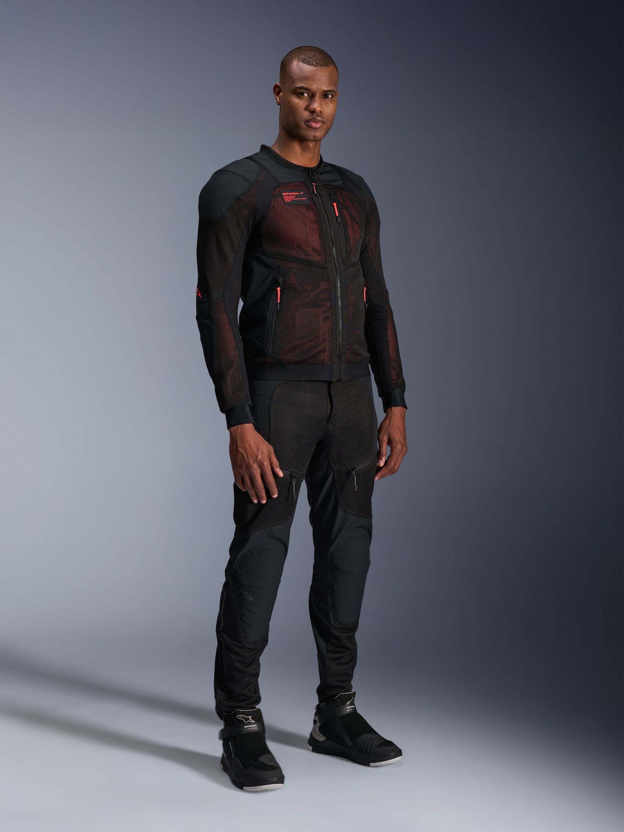 Alpinestars Troop-Air Pro Jacket in Black Red Fluo, technical armored mesh motorcycle jacket worn by a male model with matching black armored pants and high-top boots, full-length studio portrait.