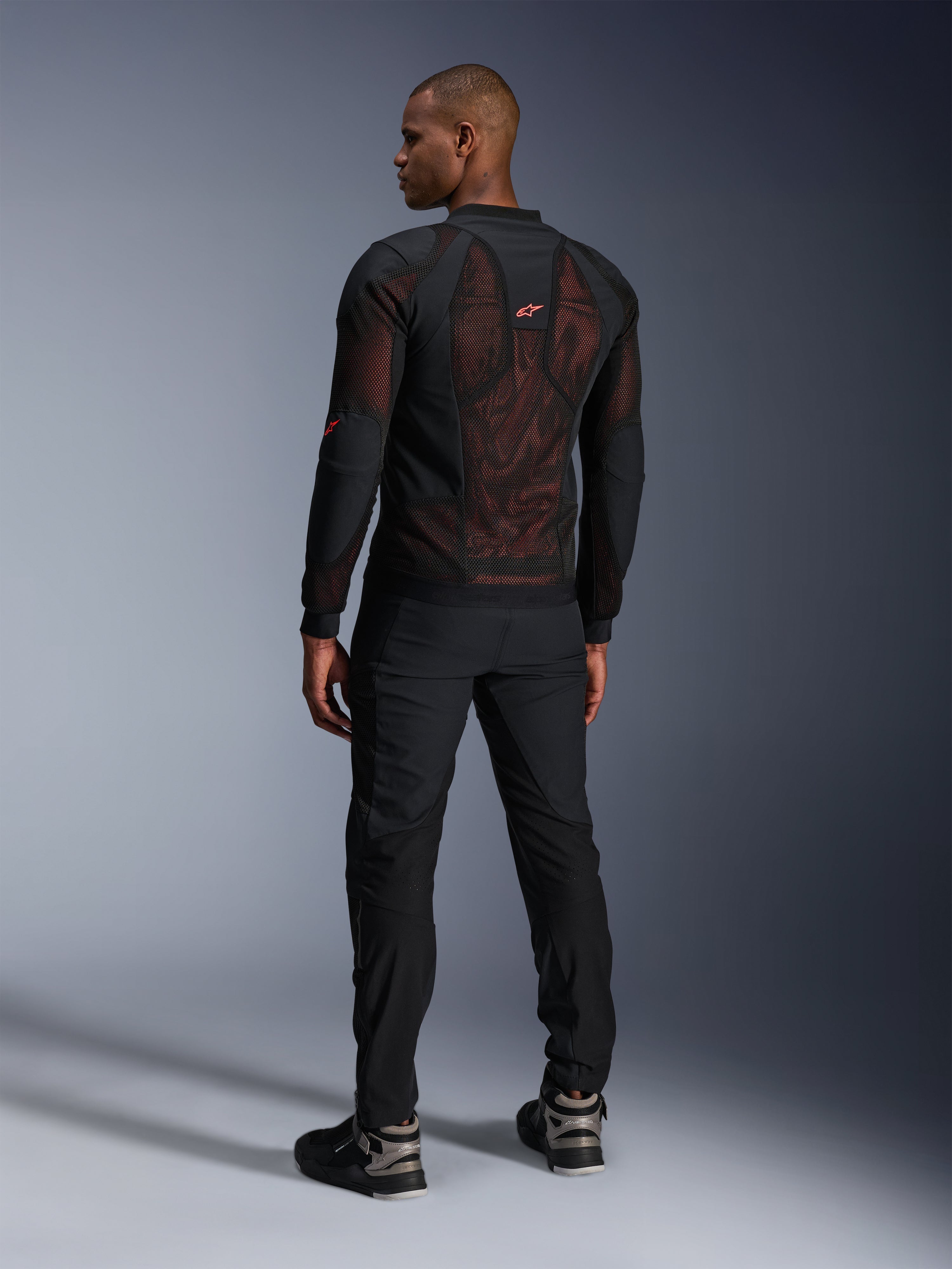 Veste Alpinestars Troop-Air Pro en Noir Rouge Fluo, vue de dos sur un mannequin, dotée d'un large panneau dorsal en mesh technique 2D avec intérieur rouge visible, associée à un pantalon de moto technique noir et des bottes noir et gris, design urbain moderne.