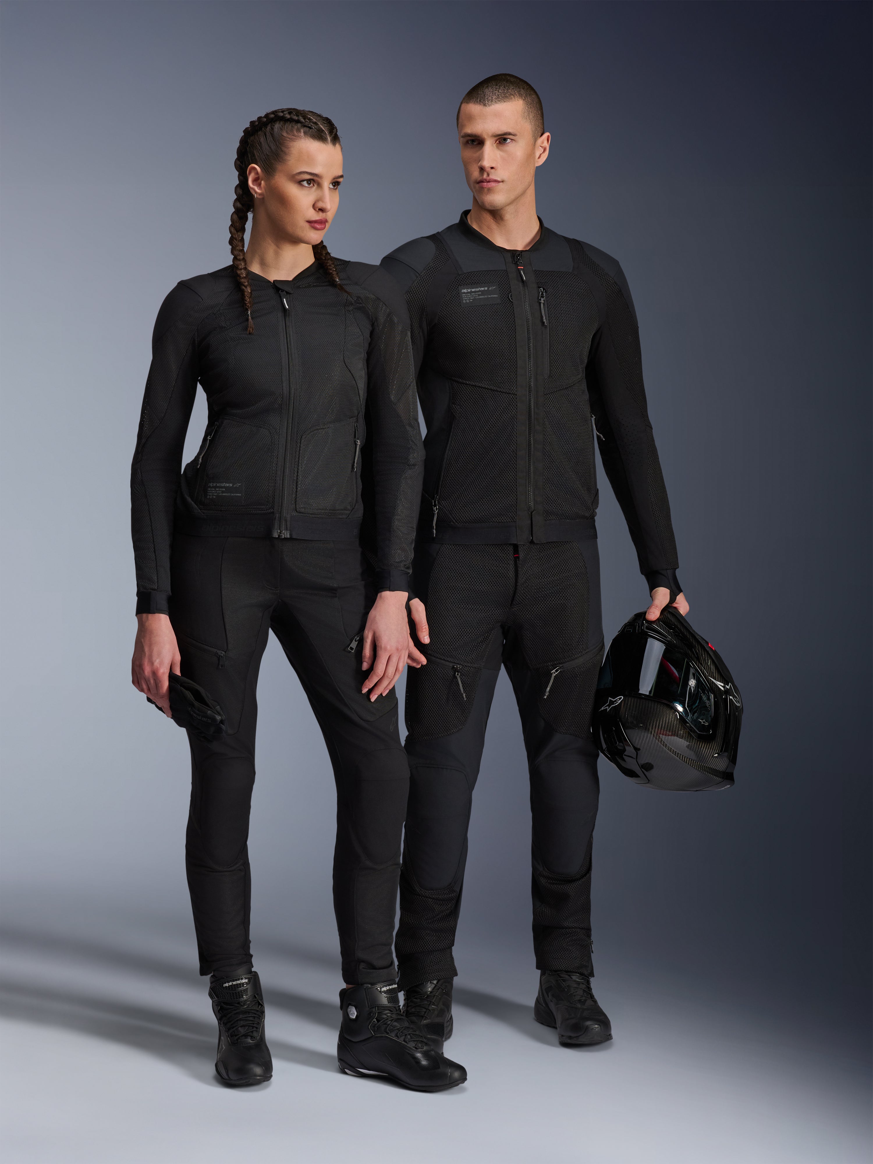Veste Alpinestars Stella Troop-Air, homme et femme portant un équipement de moto technique assorti tout en noir, doté d'une construction en maille pour la circulation de l'air, pantalons et bottes noirs, homme tenant un casque noir, tenue de moto urbaine