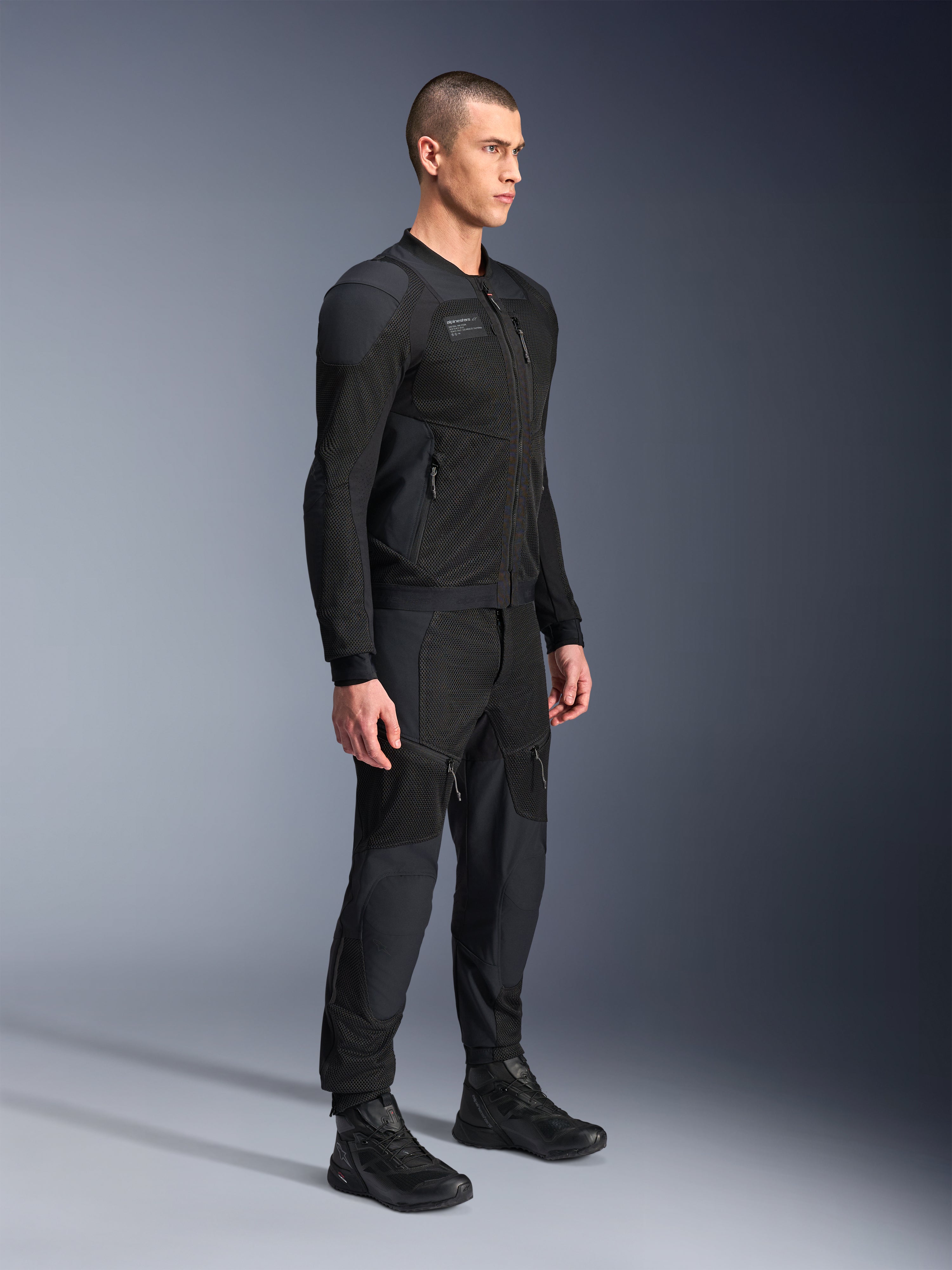 Veste Alpinestars Troop-Air Pro, veste technique ventilée et blindée, noire, portée par un mannequin avec un pantalon de moto et des bottes noirs assortis, dotée d'une structure technique en maille 2D avancée et de panneaux de ventilation respirants, équipement de conduite urbain contemporain.