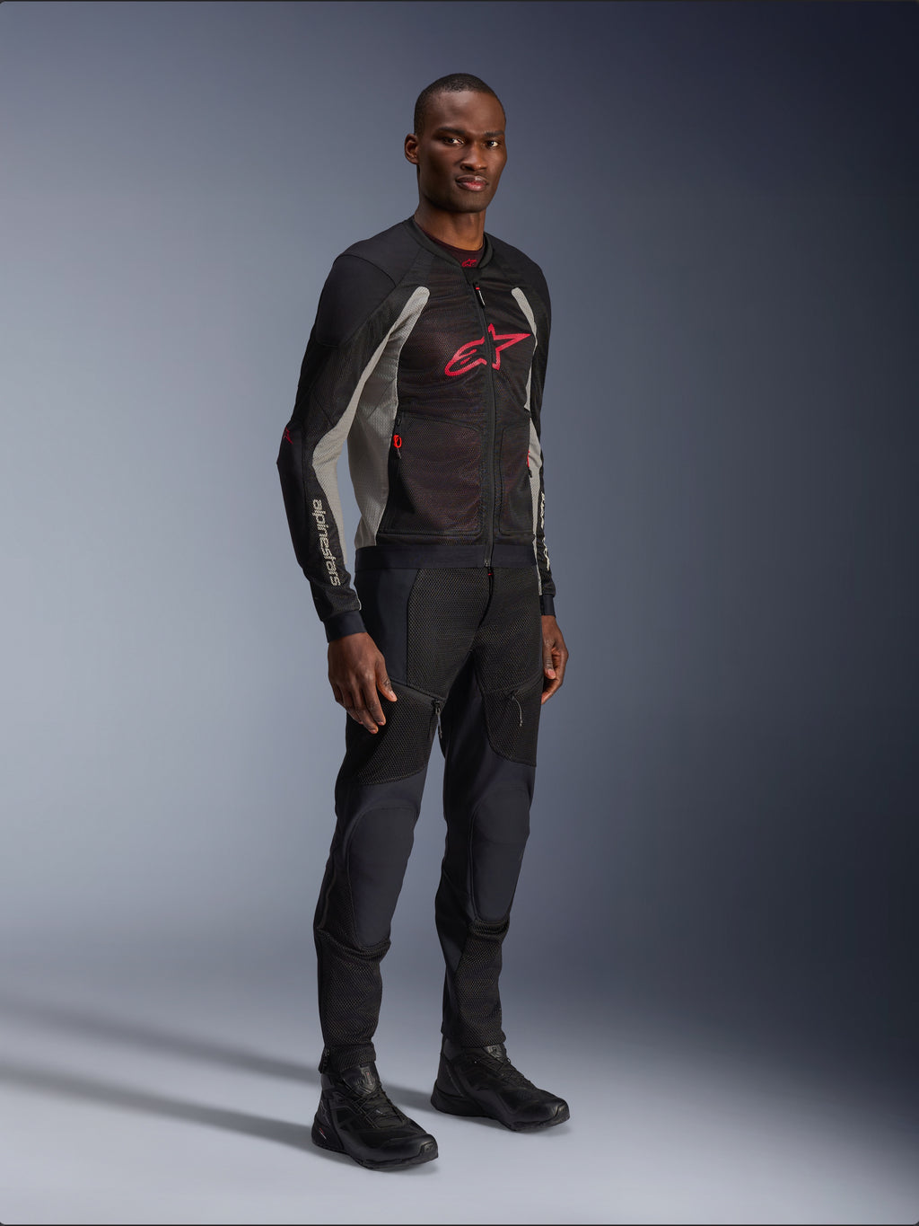 Veste Alpinestars Troop-Air, veste de moto en maille technique noir, gris cendré et rouge vif, portée par un mannequin avec pantalon et bottes noirs assortis, arborant un logo Alpinestars rouge sur la poitrine, équipement de conduite urbain contemporain