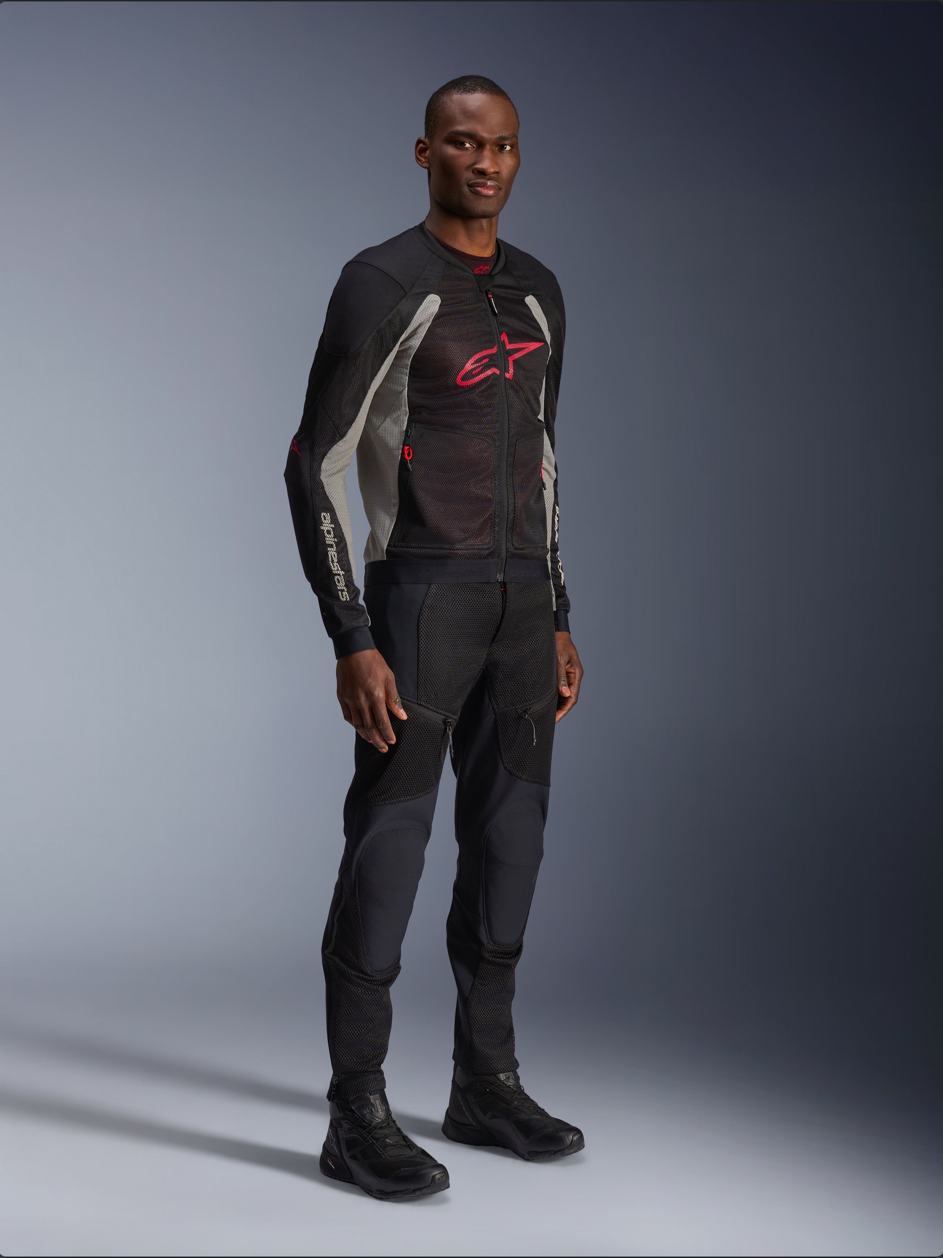 Veste Alpinestars Troop-Air, veste de moto en maille technique noir, gris cendré et rouge vif, portée par un mannequin avec pantalon et bottes noirs assortis, arborant un logo Alpinestars rouge sur la poitrine, équipement de conduite urbain contemporain