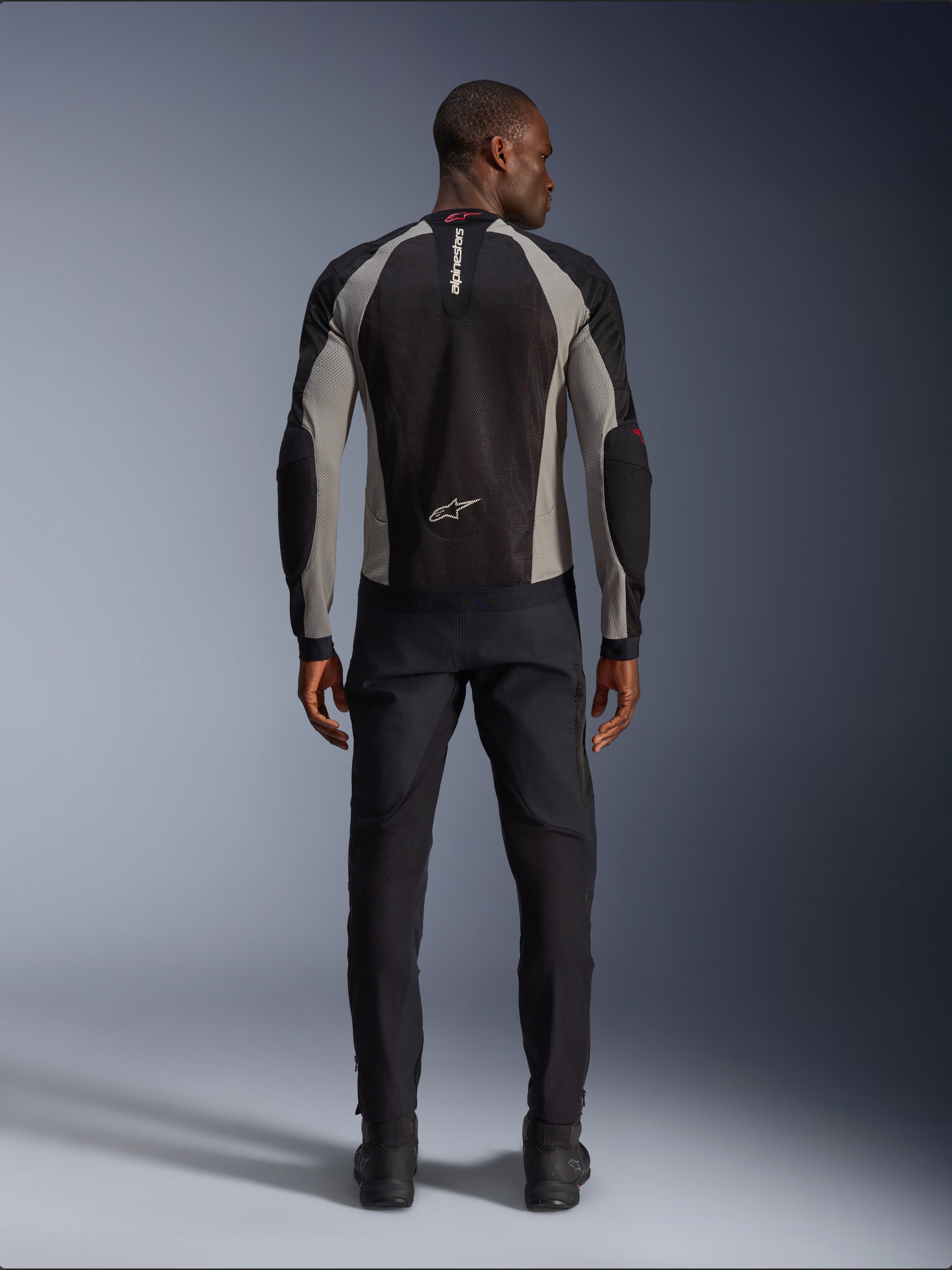 Veste Alpinestars Troop-Air, veste de moto noir, gris cendré et rouge vif, vue arrière sur un modèle masculin, construction en maille légère pour la ventilation, logos Alpinestars sur le col et le bas du dos, portée avec un pantalon de moto noir
