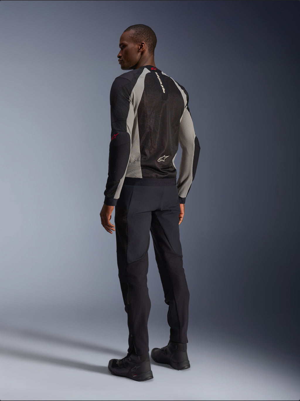 Veste Alpinestars Troop-Air vue de dos, coloris noir, gris cendre et rouge vif, construction technique en maille pour la ventilation, associée à un pantalon de moto noir et des bottes de pilotage, style urbain contemporain