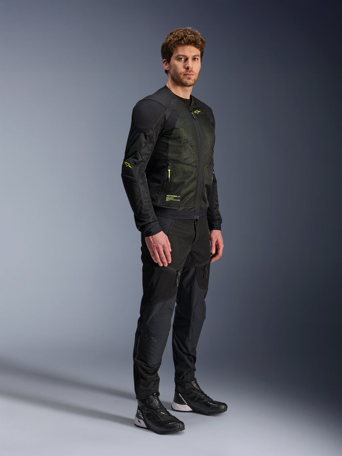 Veste Alpinestars Troop-Air, veste de moto en mesh, noir et jaune fluo, portée par un mannequin avec un pantalon noir assorti et des chaussures de moto, dotée d'une construction en mesh à haut flux d'air avec des accents jaune fluo sur la poitrine et les manches, design technique léger pour la conduite sur route
