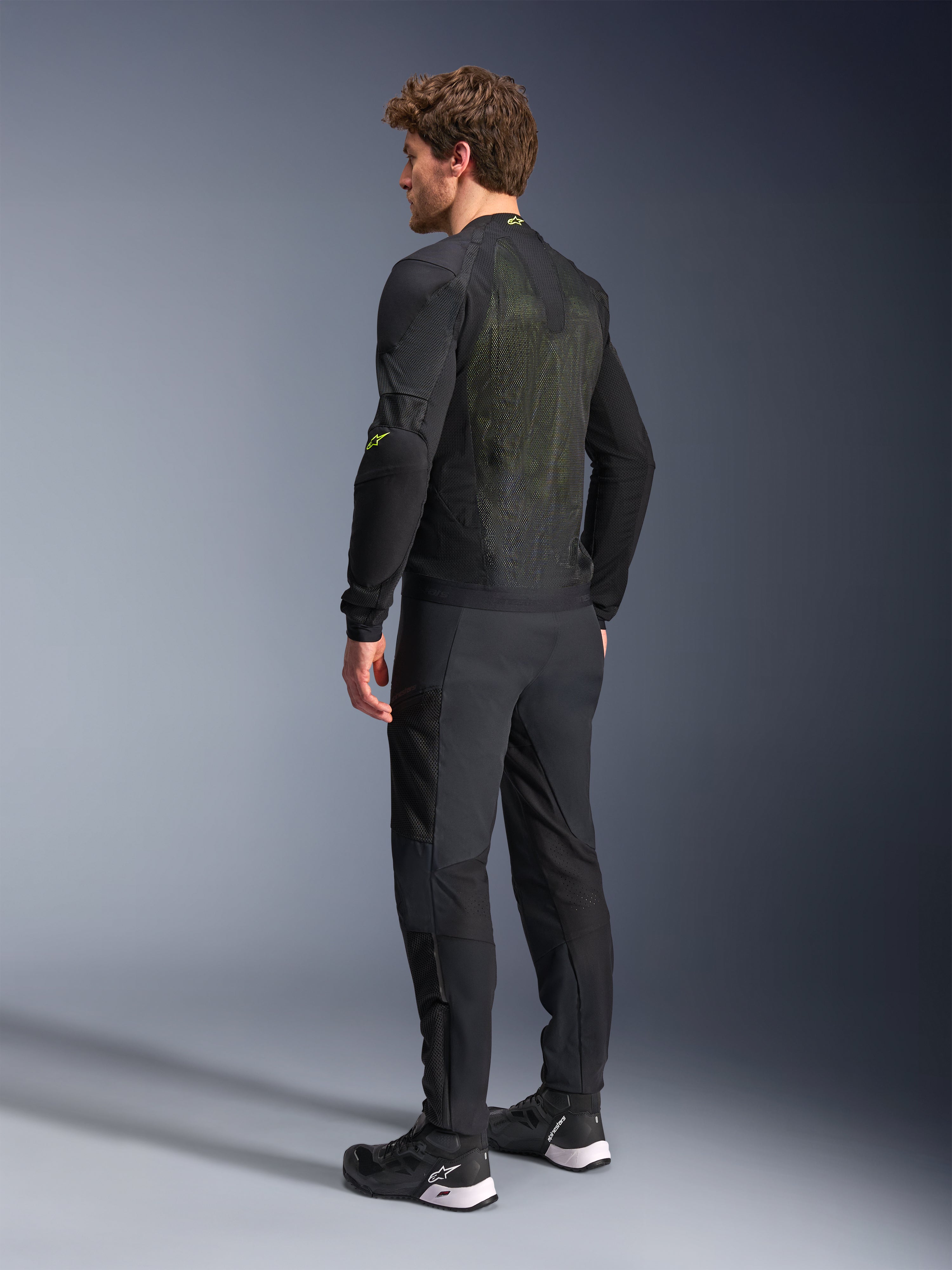 Alpinestars Troop-Air Jacket in Black and Yellow Fluo, rear view of a male model wearing the technical air jacket, featuring a large mesh back panel with yellow highlights and fluo yellow Alpinestars logos, paired with black motorcycle pants.