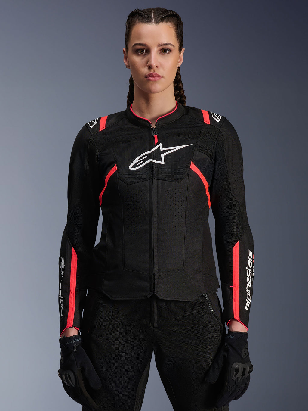 Veste Alpinestars Stella T-Sps Air V2 pour femme, noir et rose Diva Pink, veste de moto en textile dotée de panneaux en maille mesh respirante et de touches roses, logo Alpinestars blanc sur la poitrine, portée par un modèle féminin avec pantalon et gants de moto noirs.