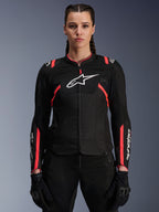 Veste Alpinestars Stella T-Sps Air V2 pour femme, noir et rose Diva Pink, veste de moto en textile dotée de panneaux en maille mesh respirante et de touches roses, logo Alpinestars blanc sur la poitrine, portée par un modèle féminin avec pantalon et gants de moto noirs.