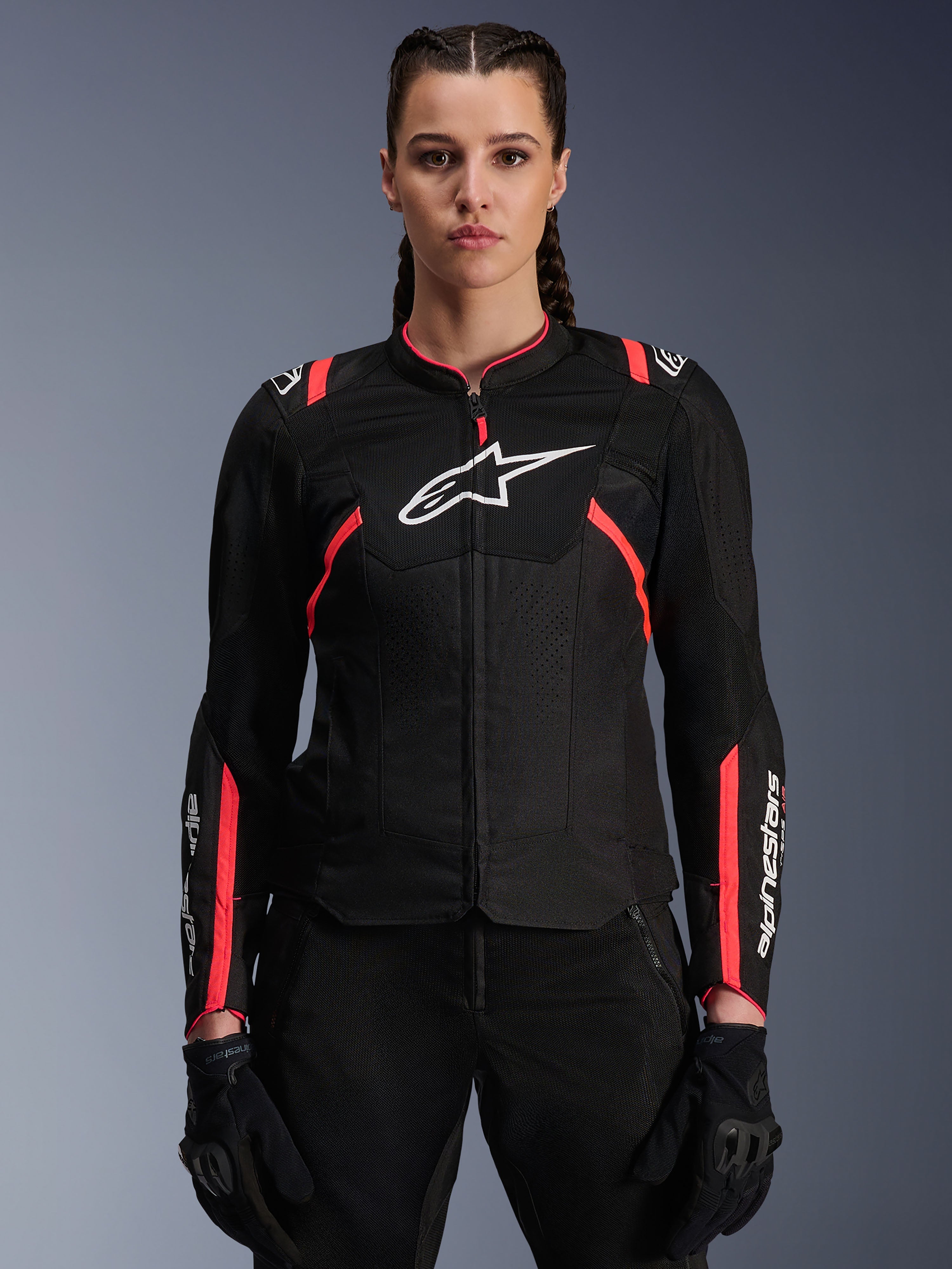 Veste Alpinestars Stella T-Sps Air V2 pour femme, noir et rose Diva Pink, veste de moto en textile dotée de panneaux en maille mesh respirante et de touches roses, logo Alpinestars blanc sur la poitrine, portée par un modèle féminin avec pantalon et gants de moto noirs.