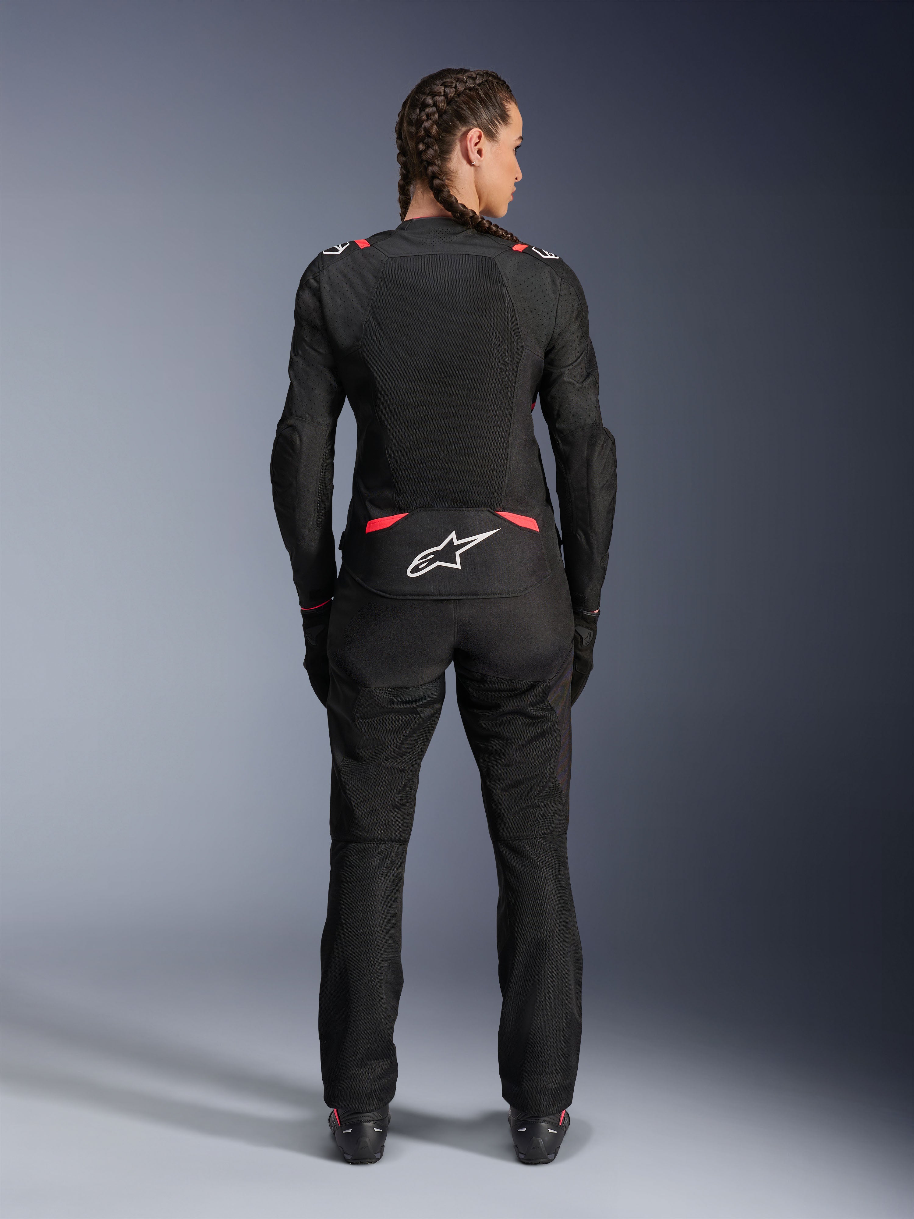 Veste Alpinestars Stella T-SPS Air V2, veste de moto en textile pour femme, noir et rose Diva, vue arrière sur un mannequin aux cheveux tressés, dotée de grands panneaux en mesh pour la ventilation, logo Astars blanc dans le bas du dos, associée à un pantalon de moto noir assorti.